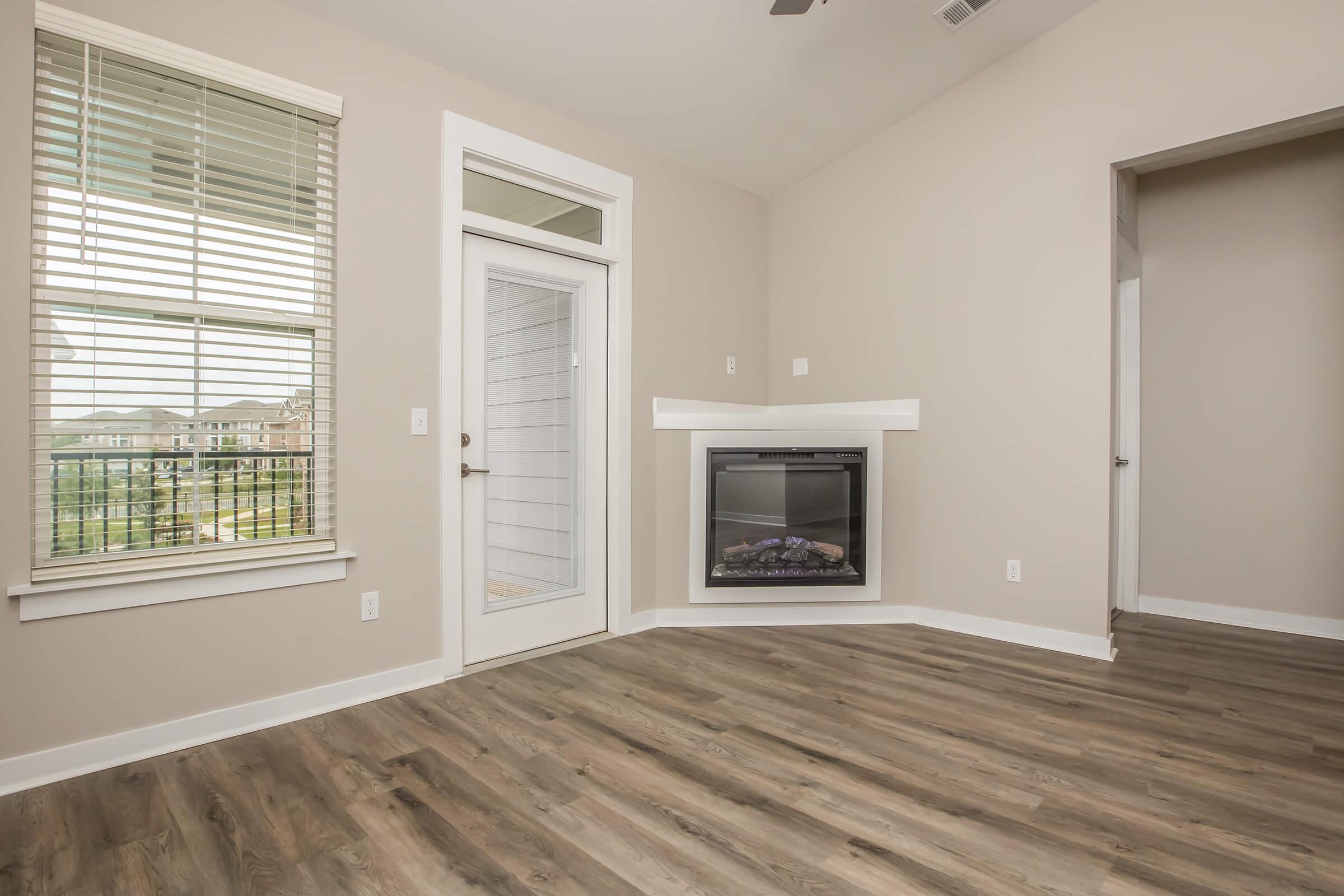 A spacious, well-lit room with large windows and neutral-colored walls. It features a modern electric fireplace and a door leading outside. The floor is covered with wood-like laminate, and the overall design is contemporary and inviting.