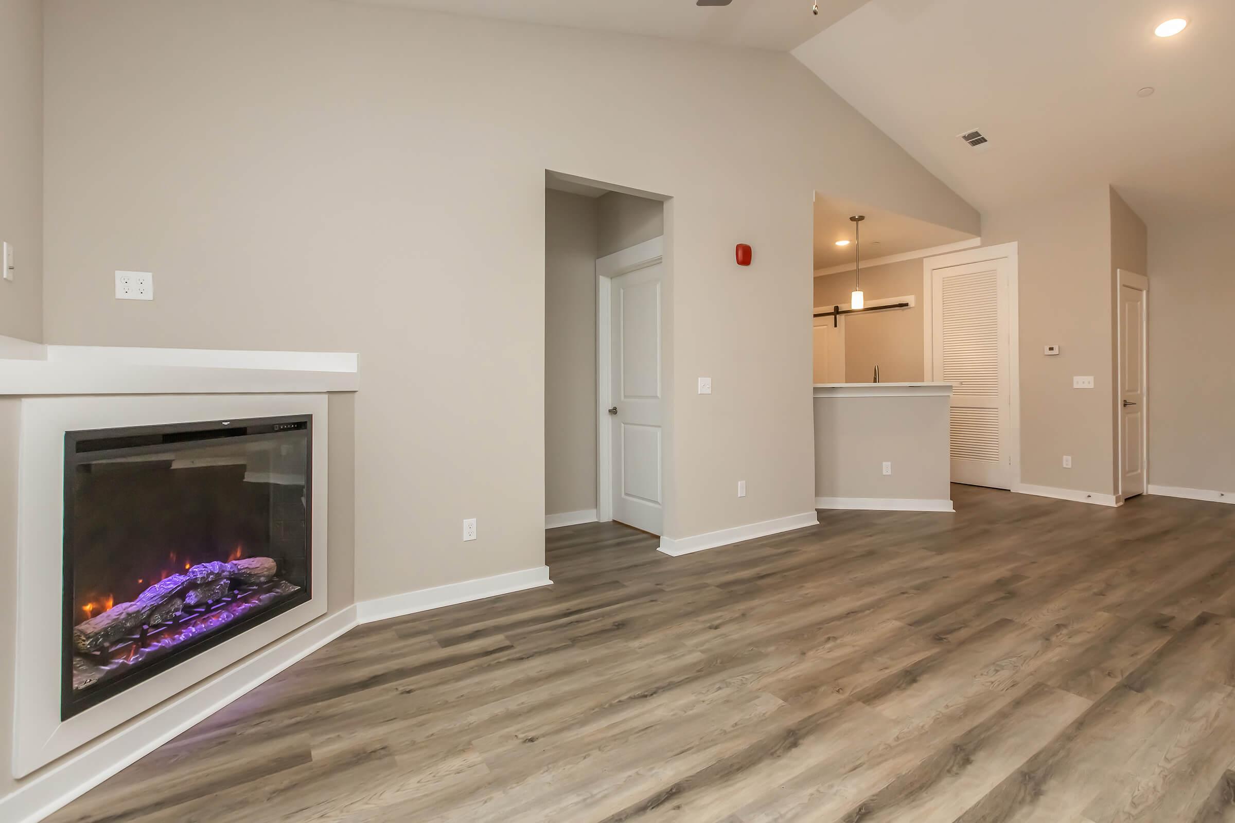 A modern living space featuring a sleek electric fireplace with purple LED accents, light-colored walls, and a wood-like laminate floor. An open doorway leads to a kitchen area with bar seating visible in the background. The room is bright and airy, emphasizing a contemporary design.