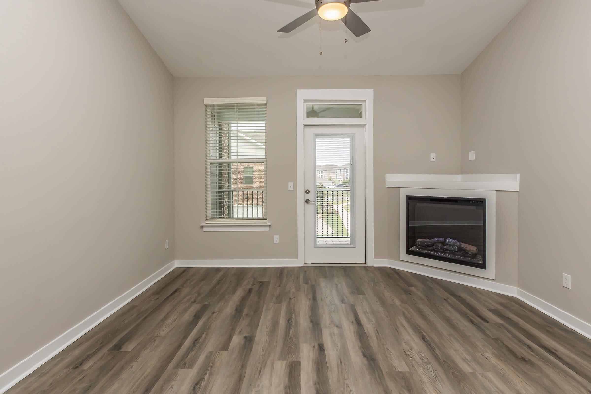 Bright and spacious living room featuring light-colored walls, a ceiling fan, and large windows. A glass door leads to a balcony, while a modern electric fireplace is situated in the corner. The floor is finished with a wood-like laminate, creating a warm and inviting atmosphere.