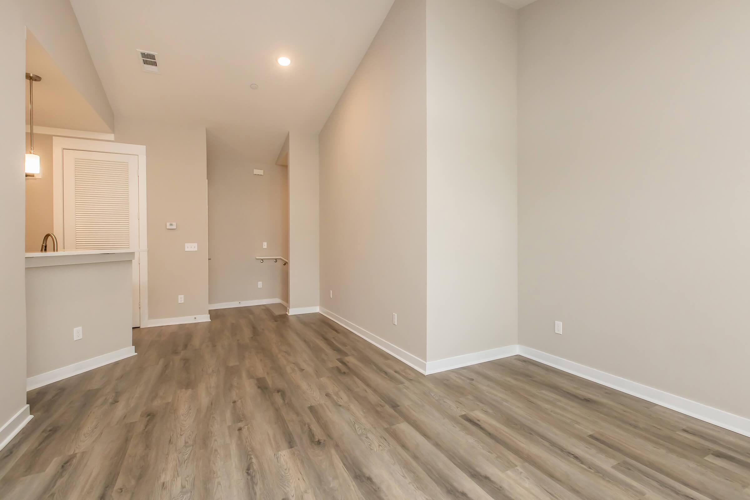 A well-lit, empty room with light gray walls and wooden flooring. The space includes a small kitchen area with a bar, a doorway leading to another area, and features minimalistic design elements. Natural light streams in, giving the room an inviting feel.