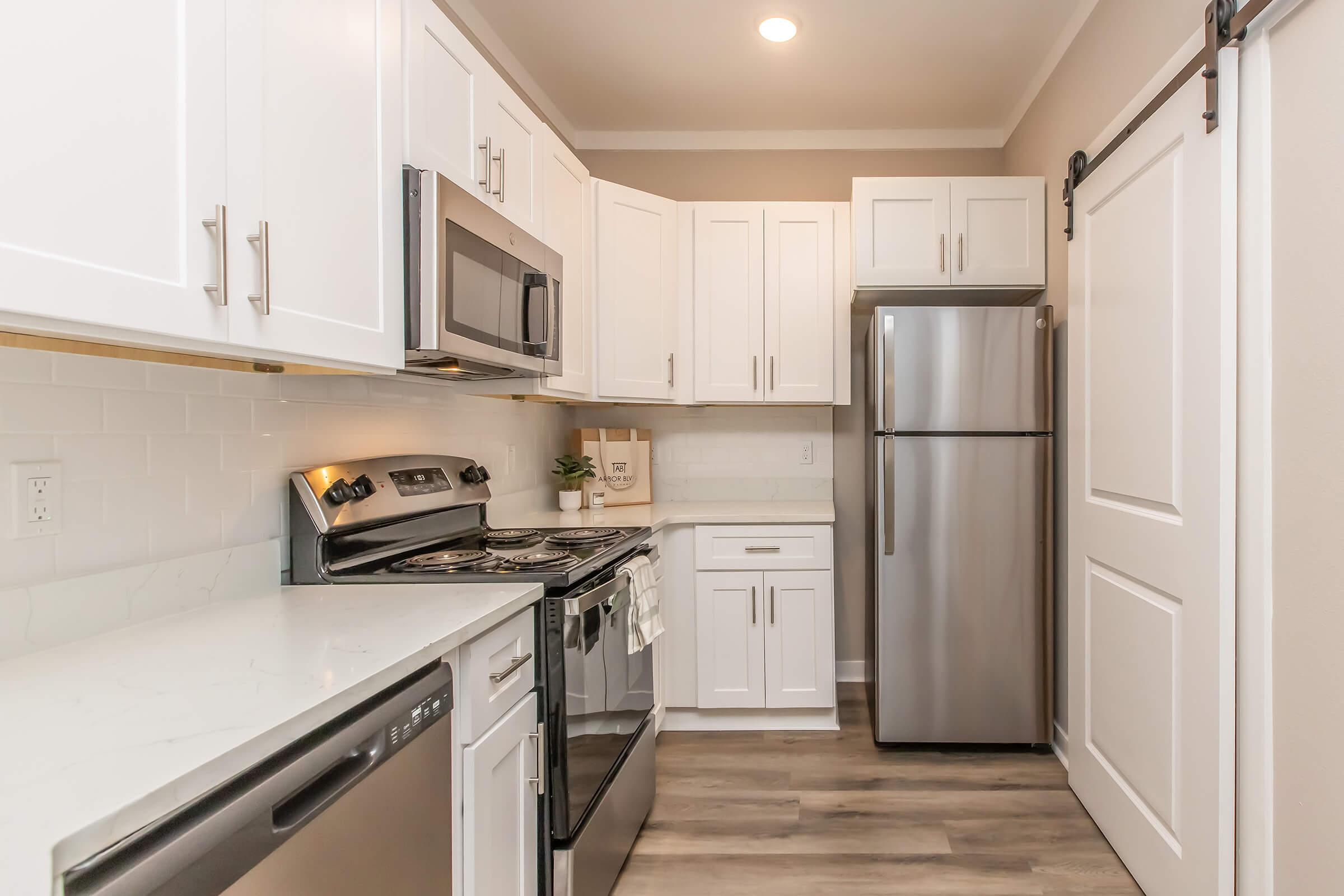 Modern kitchen featuring white cabinetry, stainless steel appliances including a microwave, oven, and refrigerator, a marble countertop, and a sliding barn door. The space is well-lit with natural light and has a clean, contemporary design.