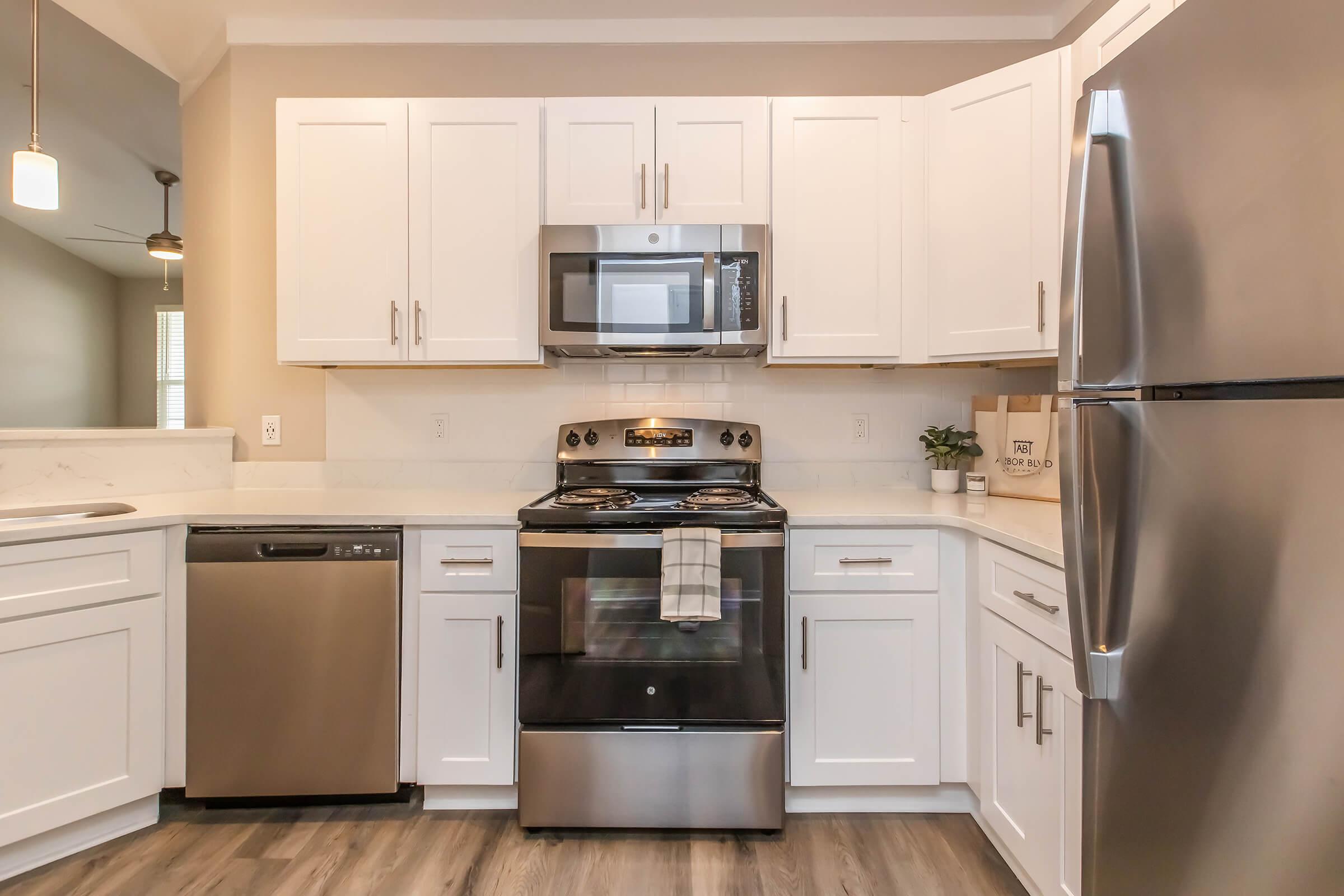 A modern kitchen featuring white cabinetry, a stainless steel refrigerator, a dishwashing machine, and an oven with a microwave above it. The countertop is light-colored, and the flooring is a wood-like laminate. There is a towel hanging on the oven, and a plant in a decorative pot on the counter.