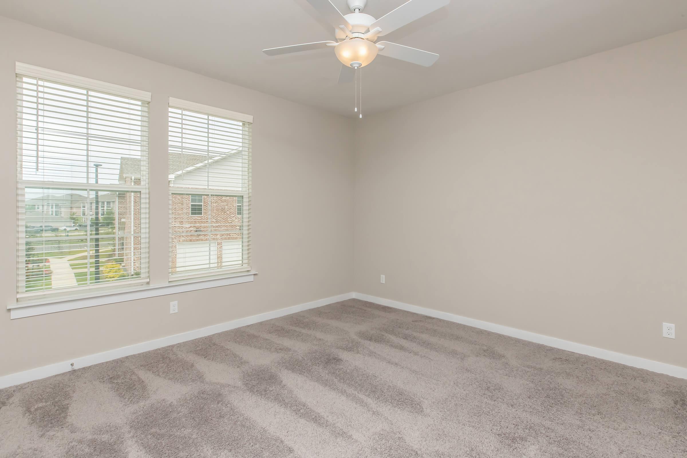 Empty room with light gray carpet and beige walls. There are two windows with blinds letting in natural light. A ceiling fan is mounted in the center of the ceiling. No furniture or decorations are present, creating a spacious and minimalist feel.