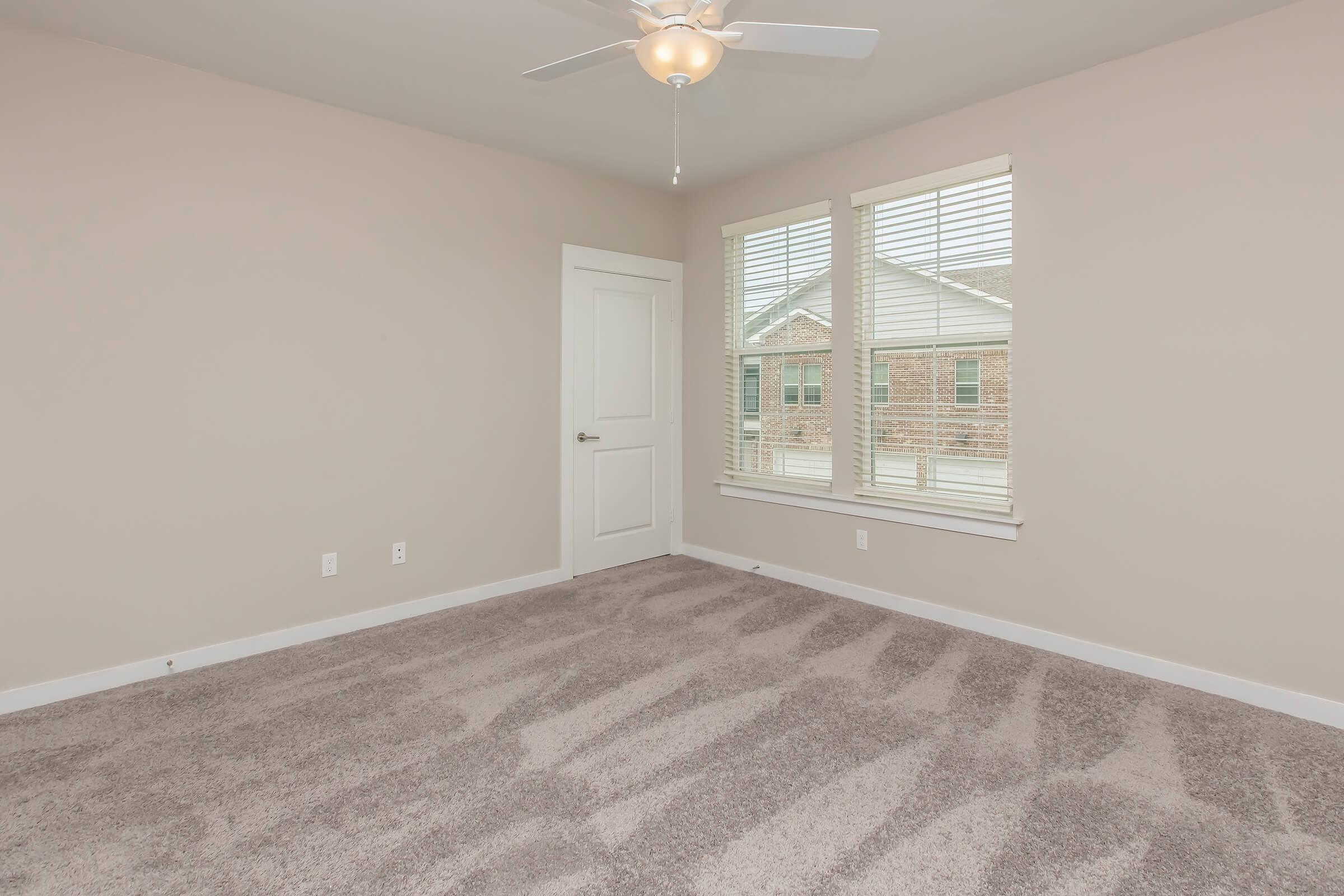 Empty room with light beige walls and carpeted floor. The space features two windows with white blinds, allowing natural light in. There is a ceiling fan and a single white door leading to another area. The overall ambiance is bright and neutral, making it suitable for various uses.
