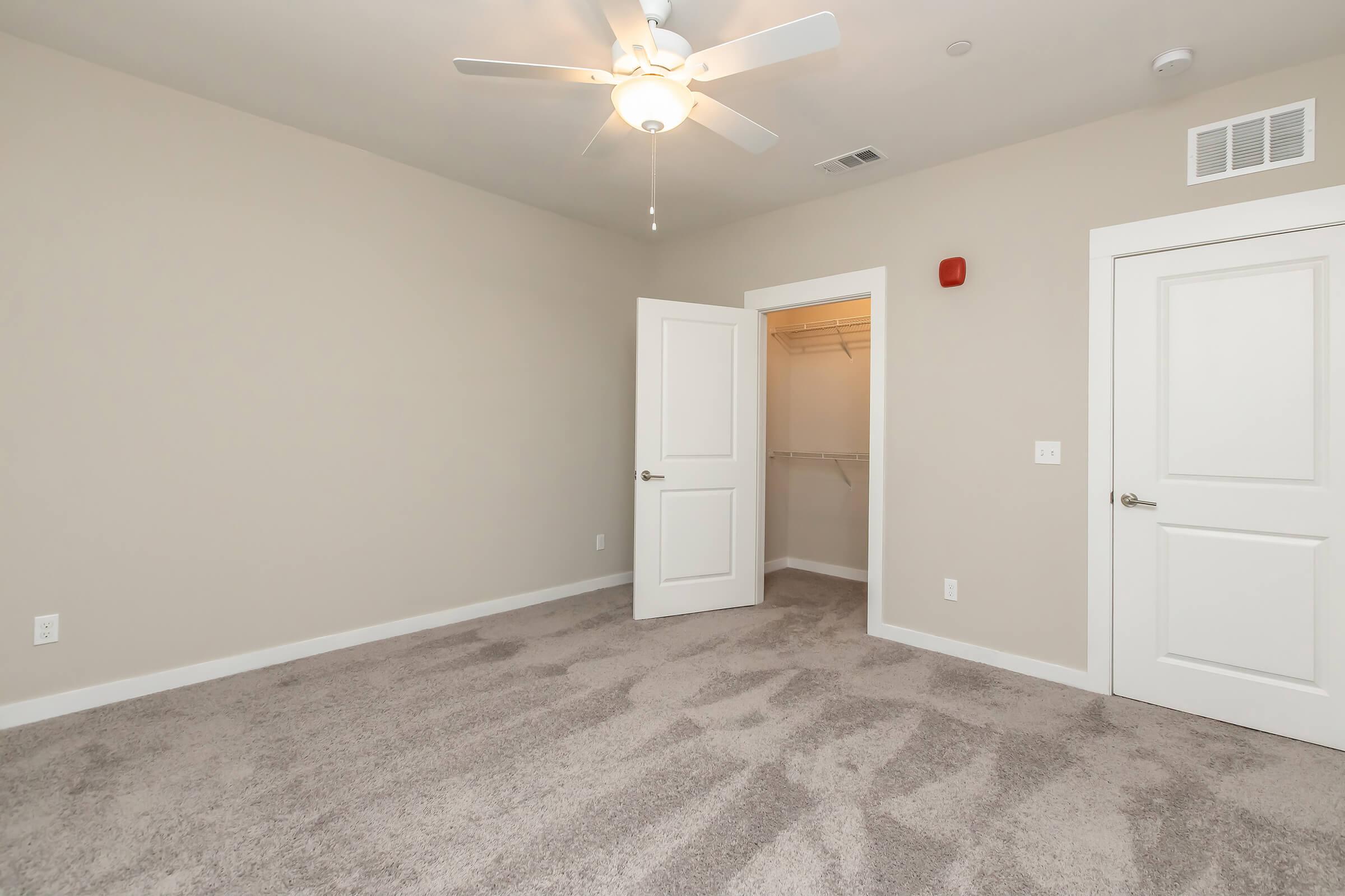 A well-lit, empty bedroom with beige walls and a ceiling fan. The floor is covered with light-colored carpet. There are two white doors; one leads to a closet, and the other opens to the room. A red alarm or sensor is mounted on the wall near the ceiling. Natural light is coming from an unseen window.
