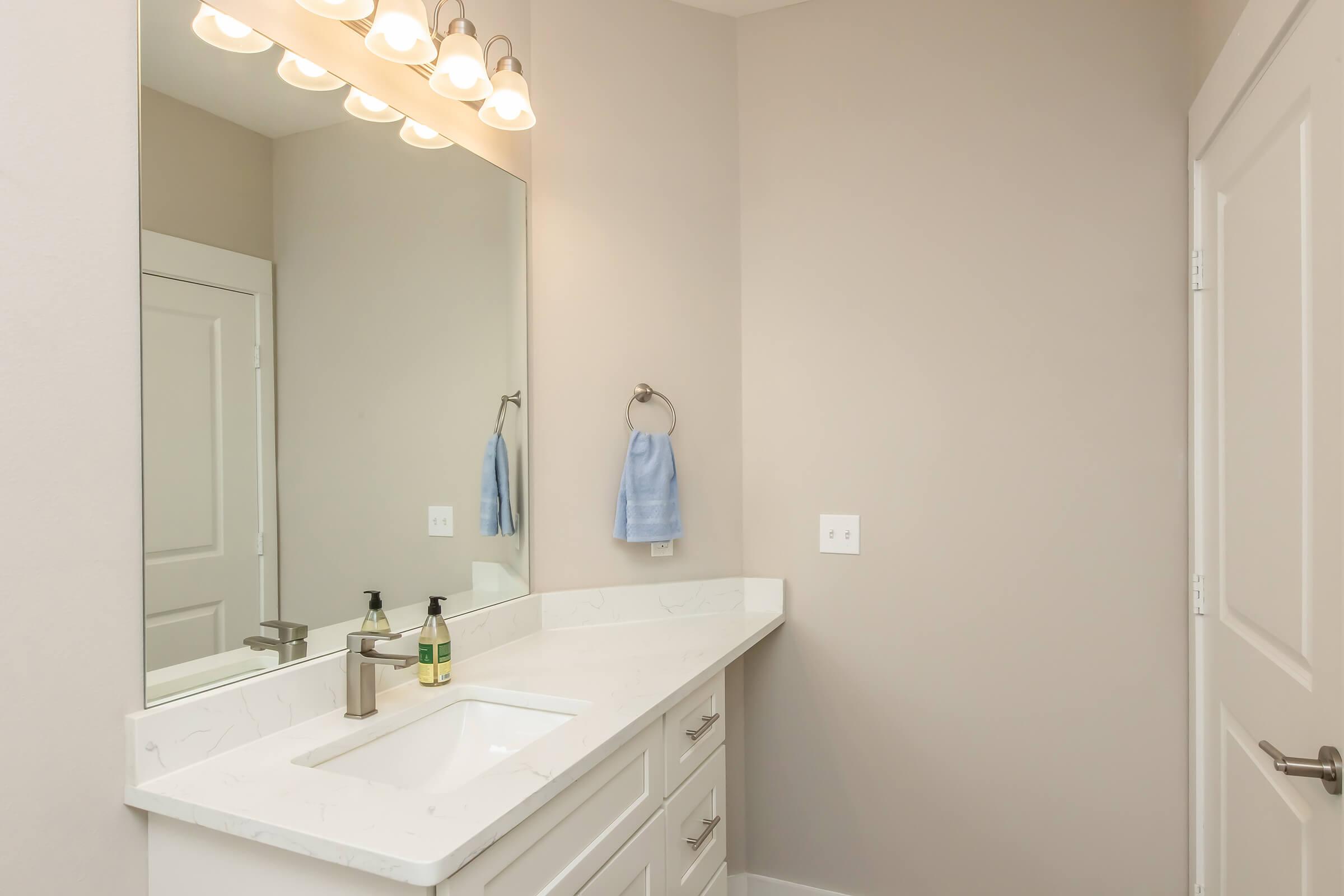 A modern bathroom featuring a white sink with a silver faucet, a large mirror above it, and two soap dispensers on the counter. The walls are a soft beige color, and there is a light fixture with multiple bulbs above the mirror. A light blue towel hangs on a towel rack nearby.