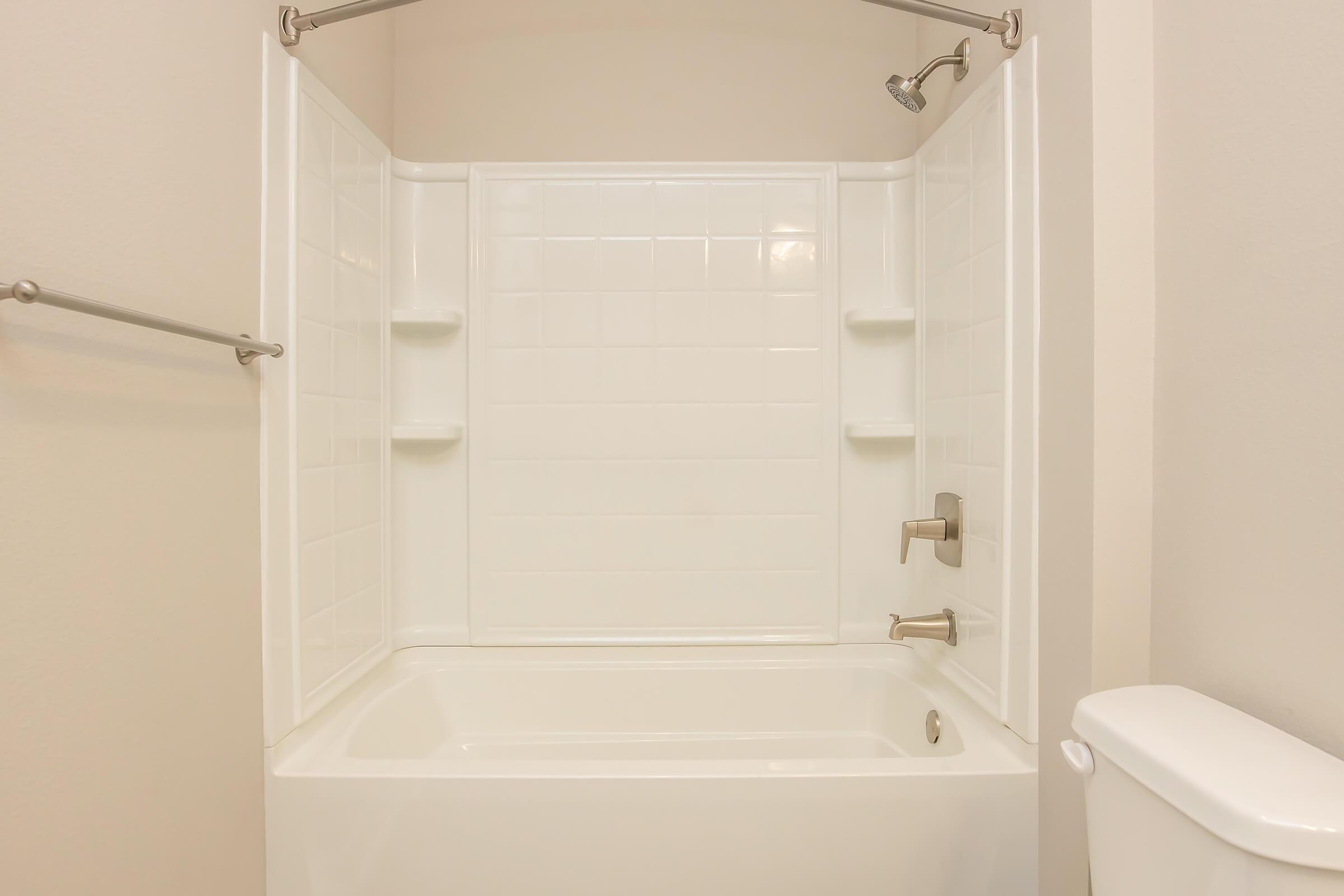 A clean, modern bathroom with a white bathtub and a shower. The walls are tiled with white square tiles, featuring built-in shelves for toiletries. A chrome showerhead and faucet are visible, along with a white toilet nearby. The overall appearance is bright and minimalist.