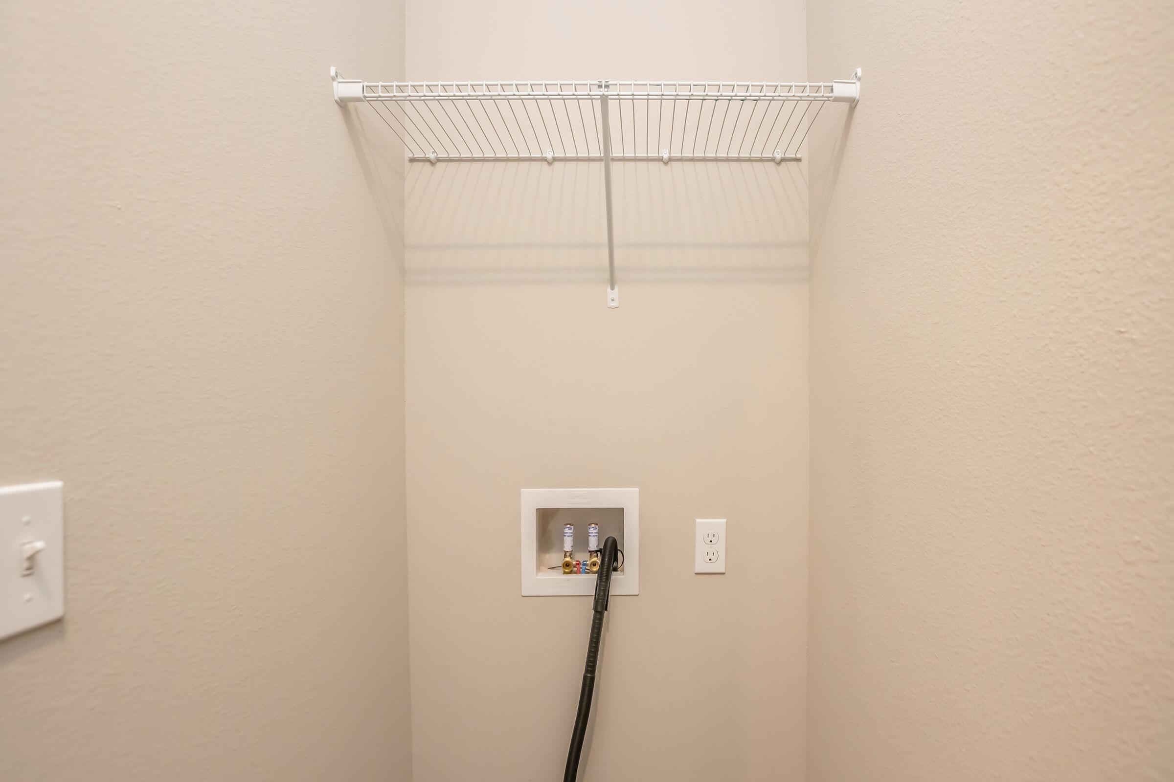 A simple laundry area featuring a blank wall painted in a light color. Above, there is a white wire shelf mounted to the wall. At the bottom, a utility box with two small bottles is visible, along with a black hose connected to the box. The space appears clean and minimalistic.
