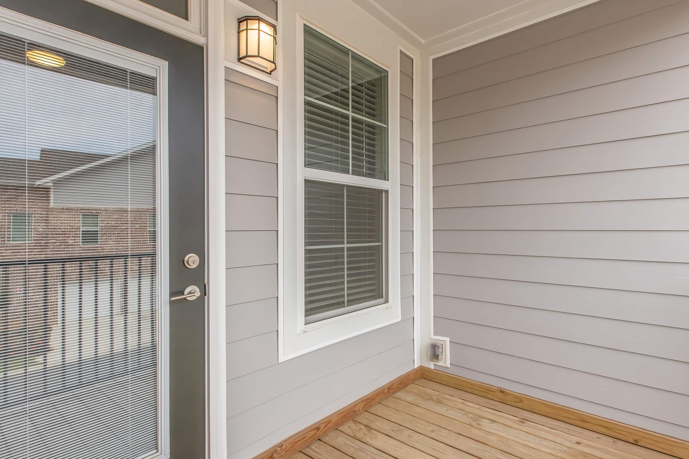 A modern porch with gray siding, featuring a glass door with a silver handle, a large window with horizontal blinds, and wooden flooring. The porch area is well-lit, with a stylish light fixture mounted near the door.