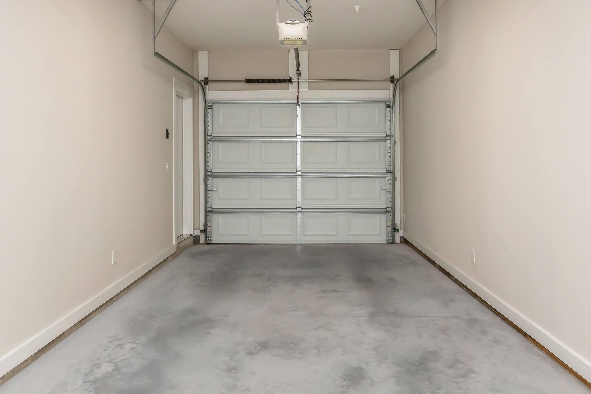 Interior of a clean, empty garage featuring a closed overhead garage door, concrete flooring, and a neutral-colored wall. There's a side door to the left, and the space is well-lit, highlighting the simplicity and spaciousness of the area.