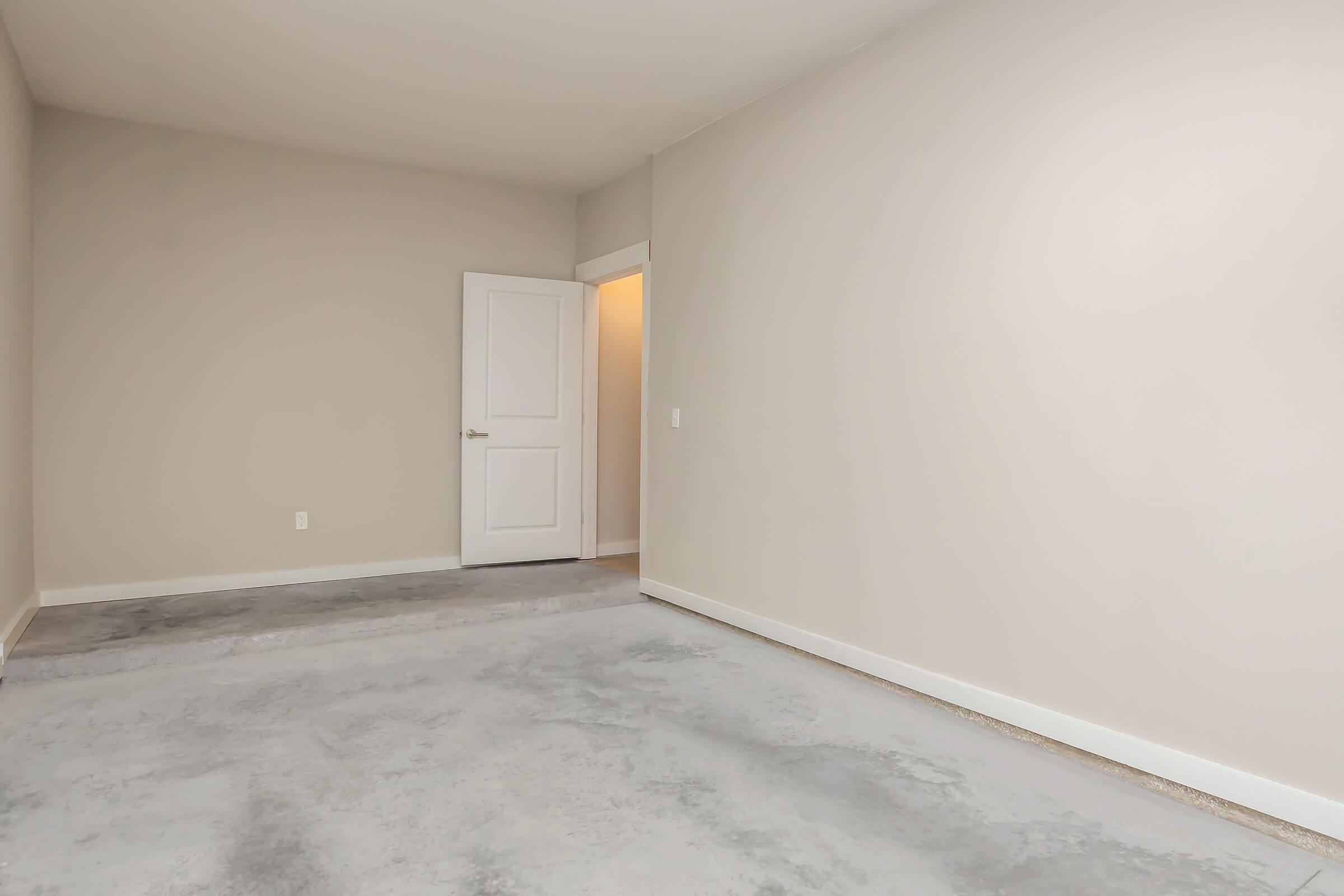 A vacant room with light beige walls and a gray concrete floor. There is a white door on the far side leading to another room, with soft lighting illuminating the space. The room is empty, creating an airy and spacious feel.