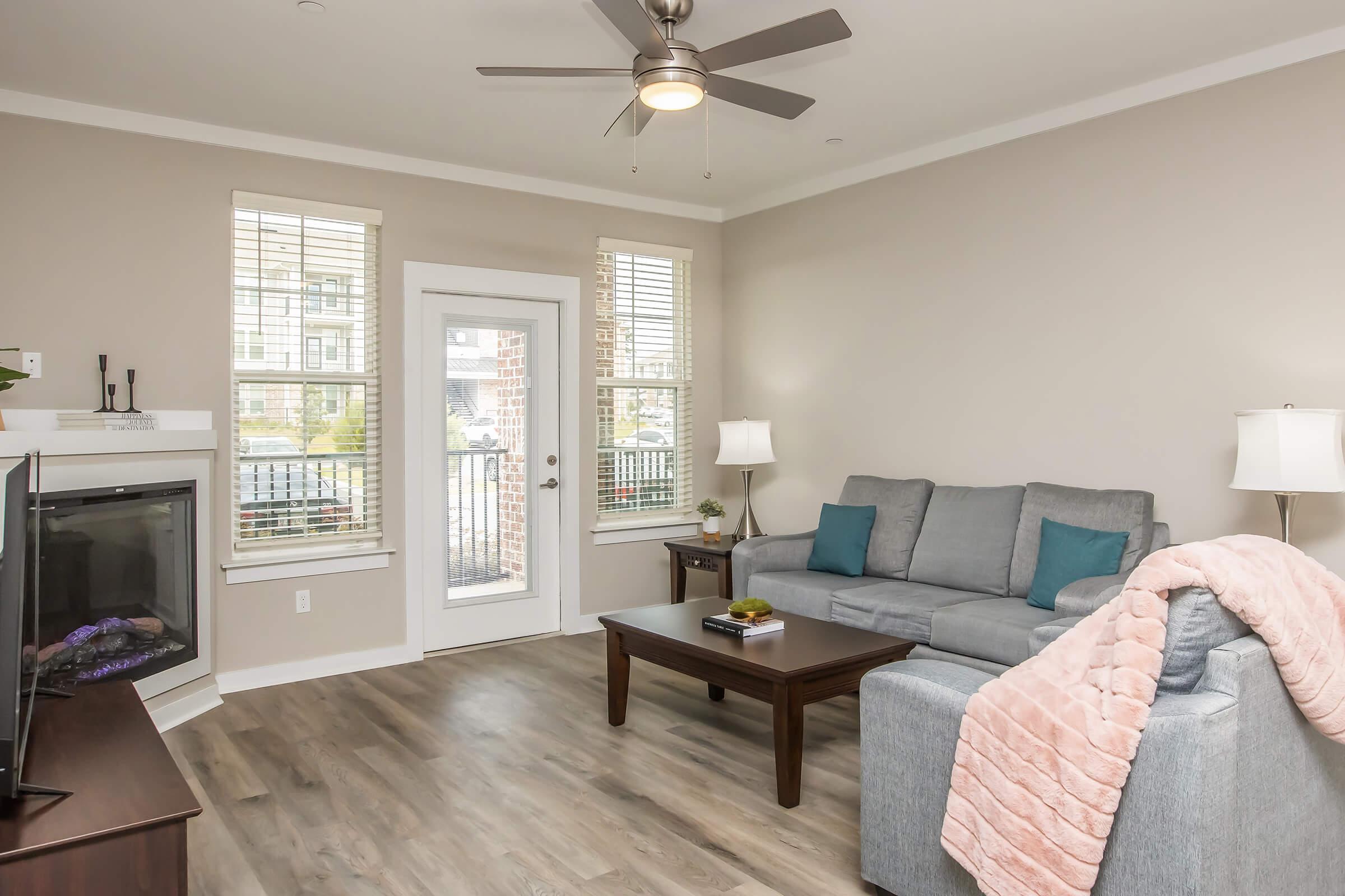 A cozy living room with a light gray sofa adorned with teal cushions, a wooden coffee table, and a TV stand. There is a soft pink throw blanket on the couch, two lamps providing warm light, a decorative plant on the table, and large windows letting in natural light. A door leads to a balcony.