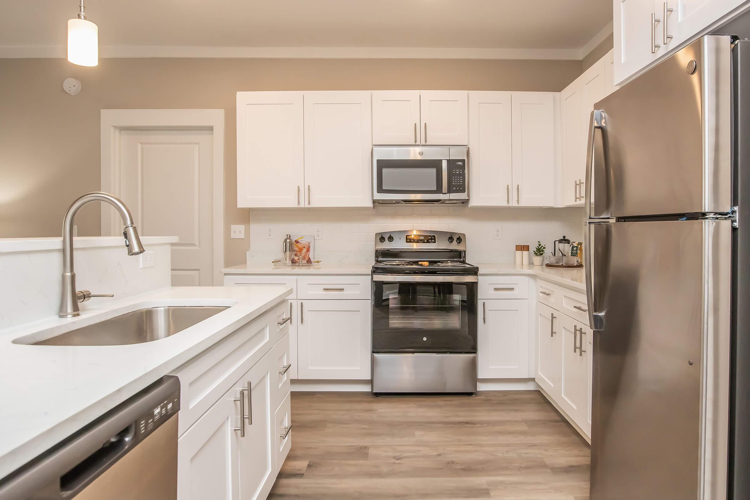 A modern kitchen featuring white cabinets, a stainless steel refrigerator, oven, and microwave. The countertops are light-colored with a farmhouse sink. The floor is a light wood finish, and the walls are a soft beige. There are decorative items on the counter and a pendant light hanging above.