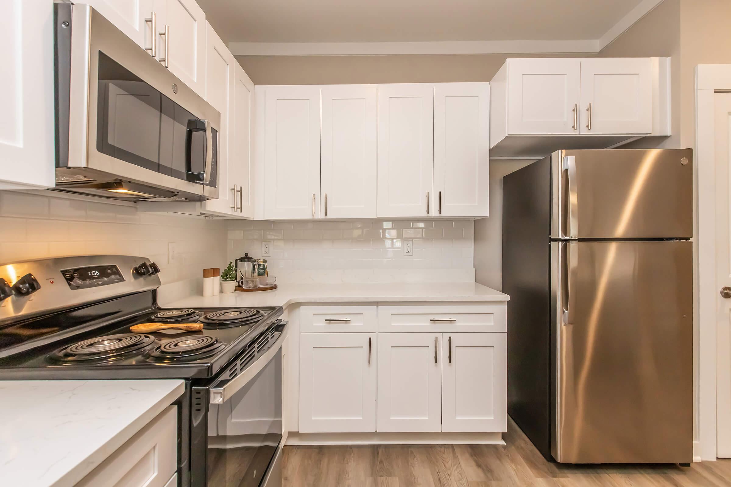 Modern kitchen with white cabinets, stainless steel appliances including a microwave and refrigerator, a black stove, and a light-colored countertop. The space features a clean, contemporary design with tile backsplash and laminate flooring.