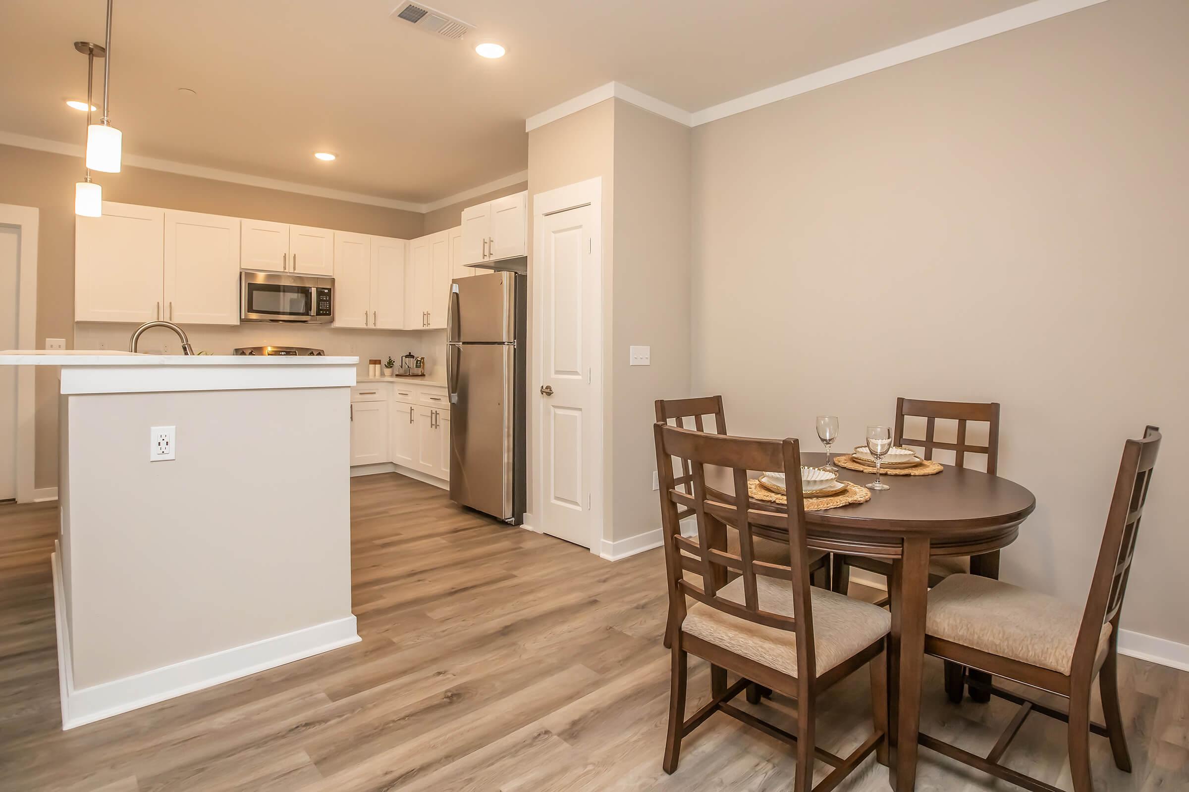 A modern kitchen and dining area featuring light wooden flooring, a round dining table with four chairs, and a kitchen with white cabinets, stainless steel appliances, and a small island. Soft lighting enhances the inviting atmosphere.