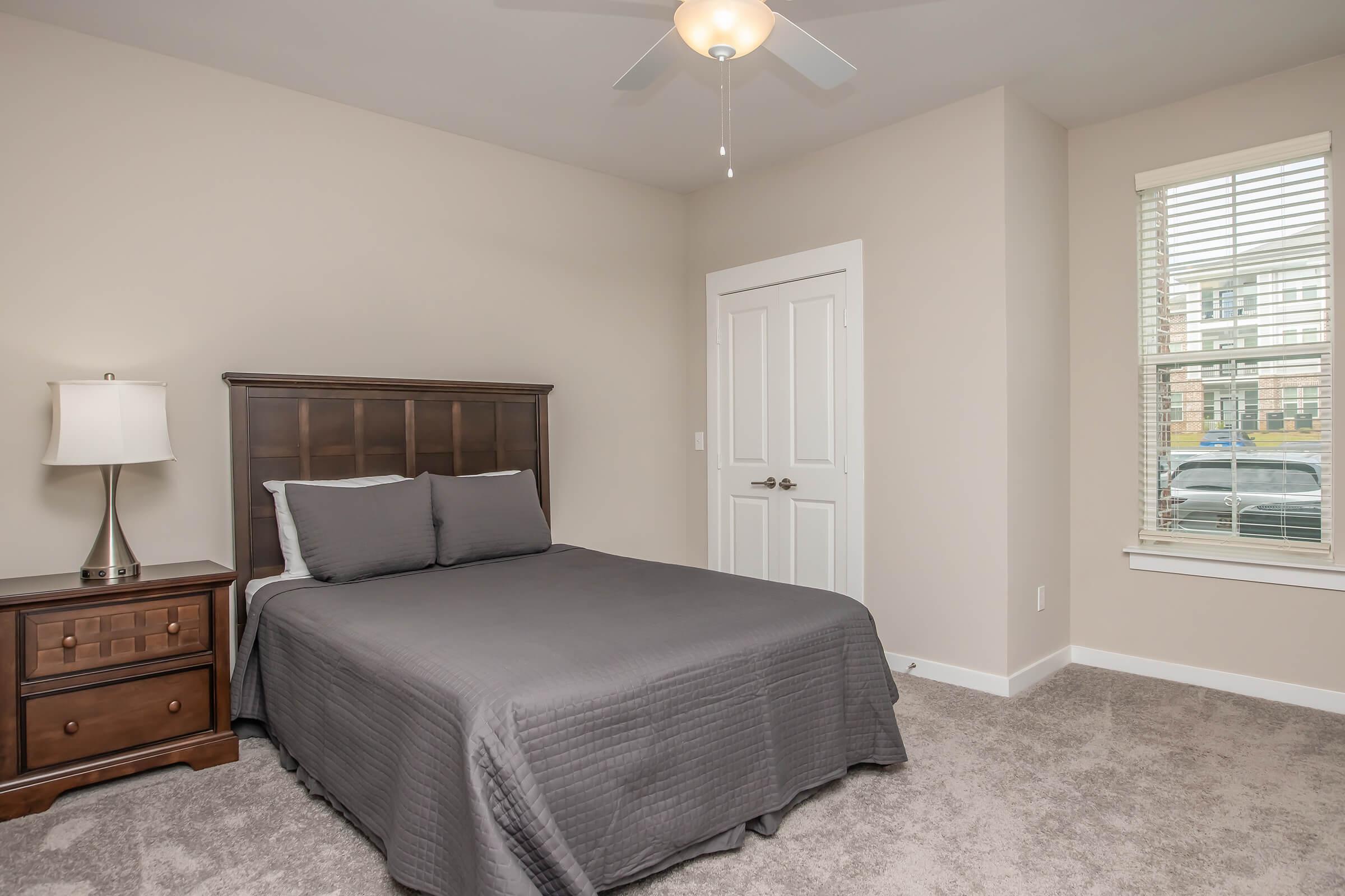 A cozy bedroom featuring a queen-sized bed with gray bedding, a wooden nightstand with a lamp, and a window with blinds allowing natural light. The walls are painted in a neutral tone, and the floor is carpeted. A door leads to another area, adding to the room's spacious feel.