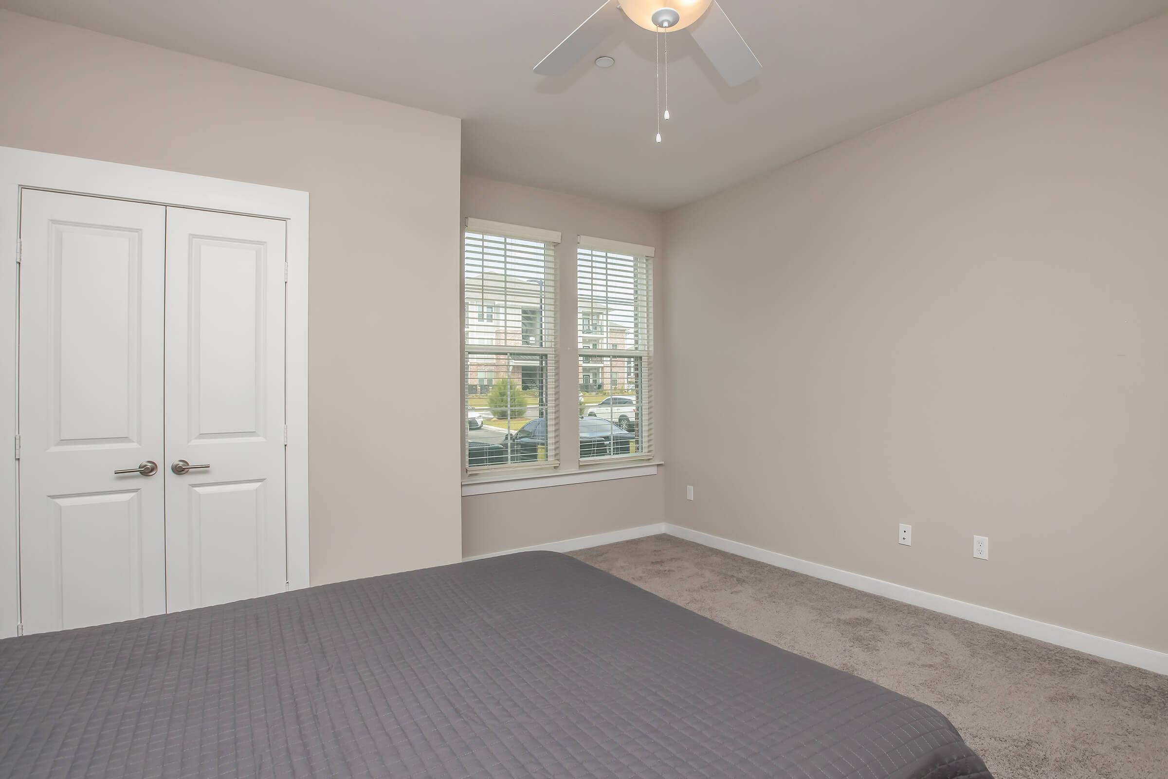 A cozy bedroom featuring light beige walls, a ceiling fan, and a large window with blinds allowing natural light. The room has a neutral-colored quilted bedspread and a plush carpet. A closed double door closet is visible on one side, with minimal decor on the walls for a clean, simple aesthetic.