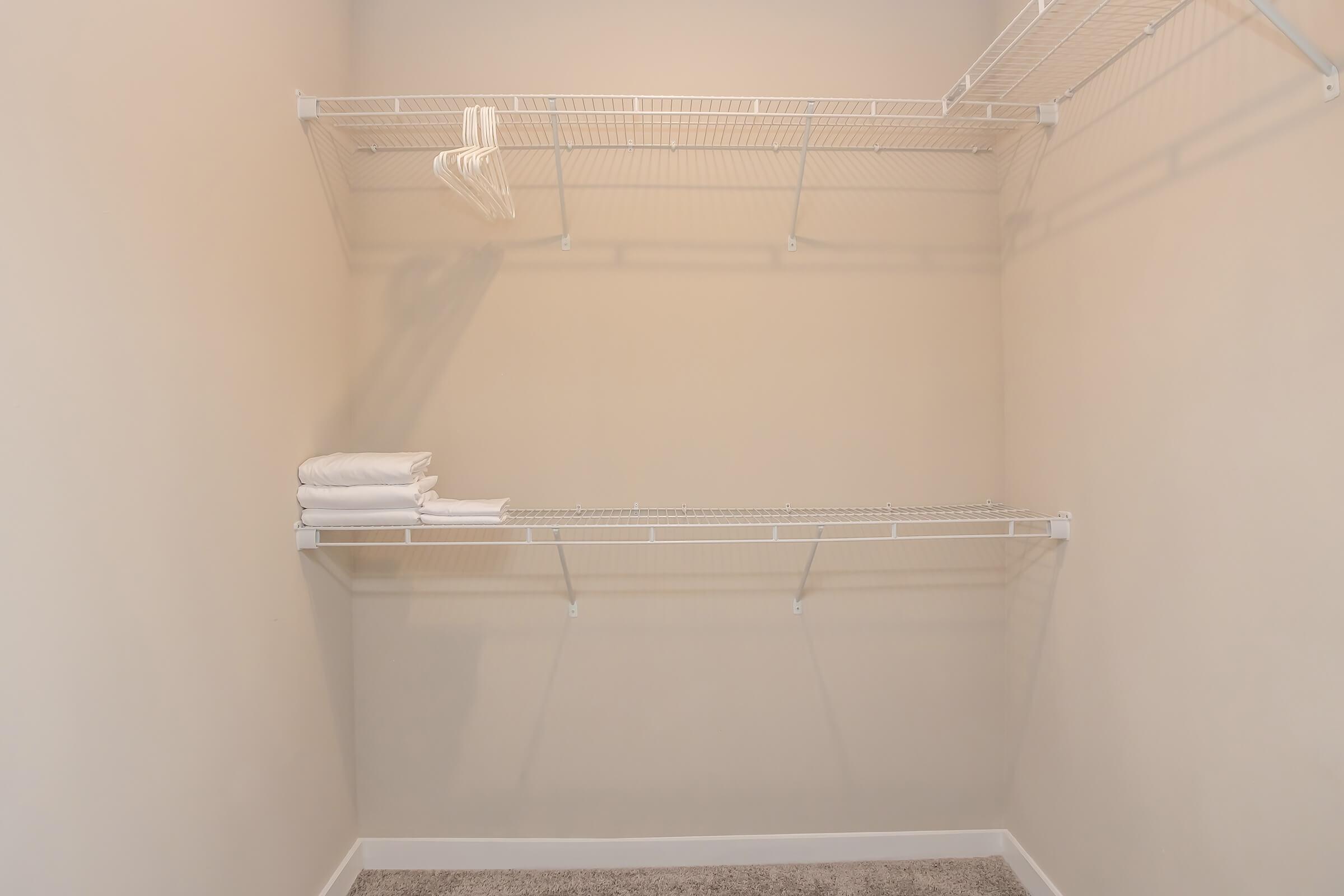 A clean, empty closet interior with beige walls. Two wire shelves are mounted on the walls, one partially filled with neatly stacked white towels. The floor is carpeted in a light color, creating a spacious and uncluttered appearance.