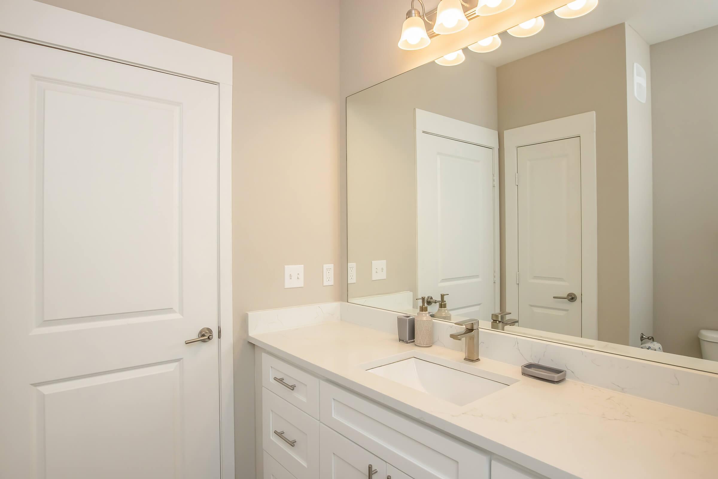 A modern bathroom featuring a white vanity with a sink, marble countertops, and a large mirror above. Soft lighting is provided by three bulbs, and the wall is painted in a neutral color. A closed door and a glimpse of a toilet can be seen in the background, creating a clean and spacious appearance.