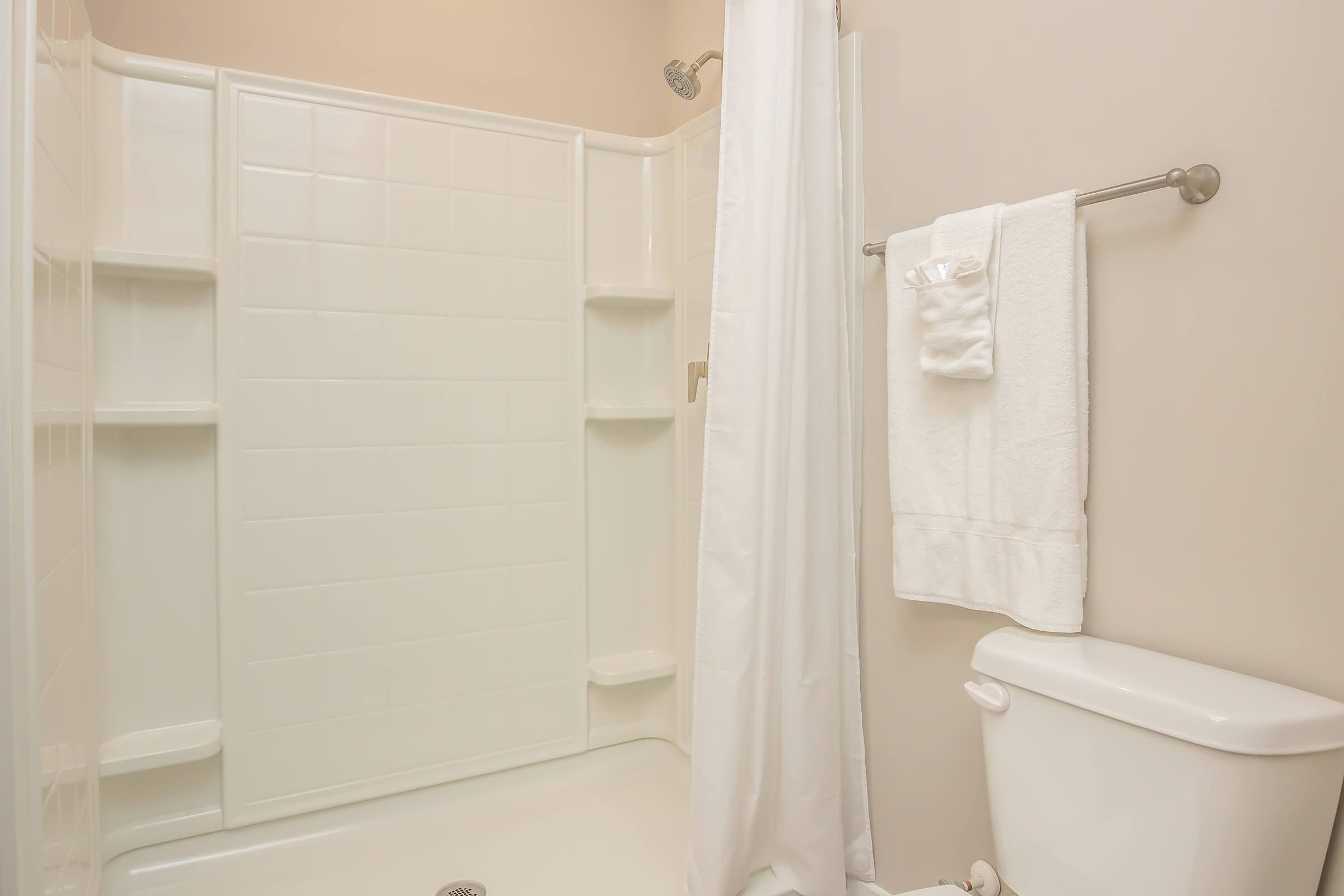 A clean and modern bathroom featuring a white shower stall with built-in shelves, a white shower curtain, and a neatly hung set of white towels. A white toilet is visible in the corner, set against a light-colored wall. The overall aesthetic is bright and uncluttered.