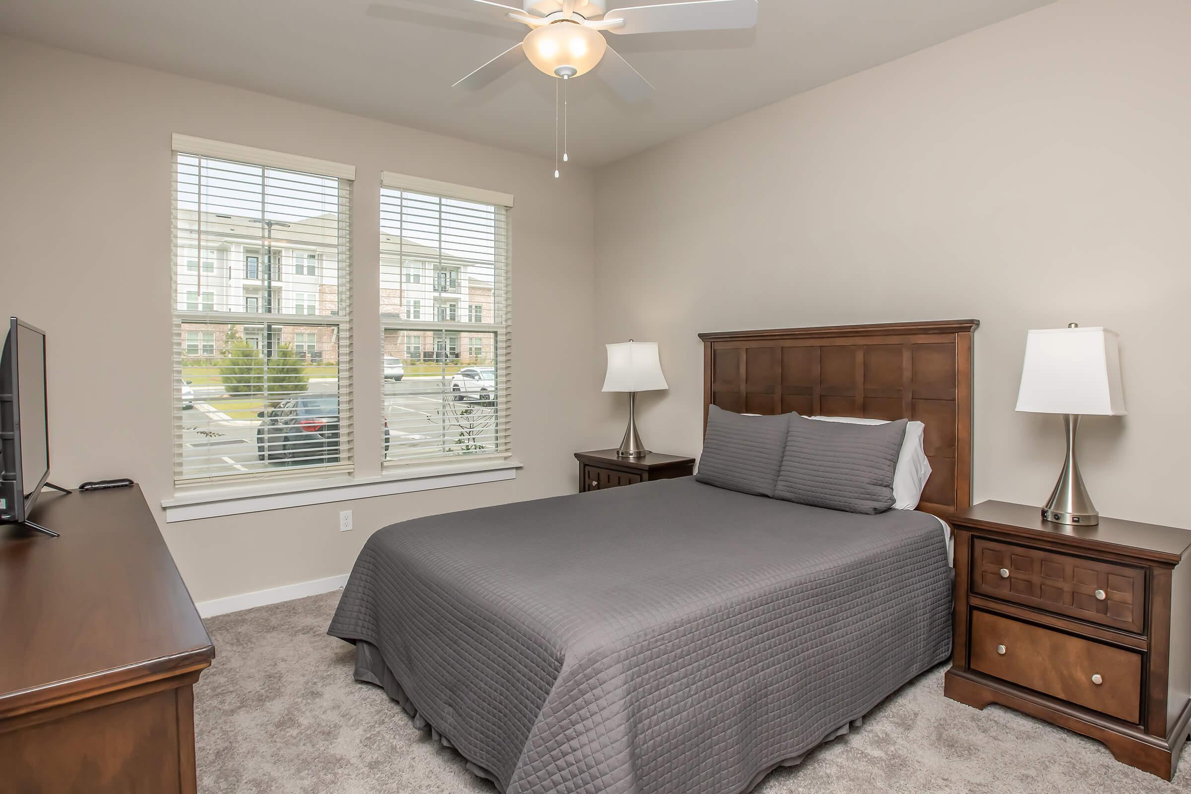 A cozy bedroom featuring a queen-sized bed with a gray quilt, wooden nightstands with lamps on either side, a dresser with a TV, and large windows showcasing a view of an outdoor area. The walls are painted a neutral color, and the carpeted floor adds warmth to the space.