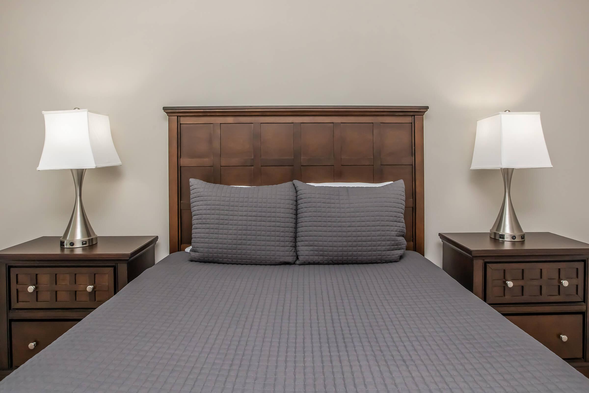 A neatly made bed with a dark gray quilt on a wooden frame, featuring two large, textured pillows. On either side of the bed, there are wooden nightstands with modern, silver lamps. The room has neutral-colored walls, creating a calm and inviting atmosphere.