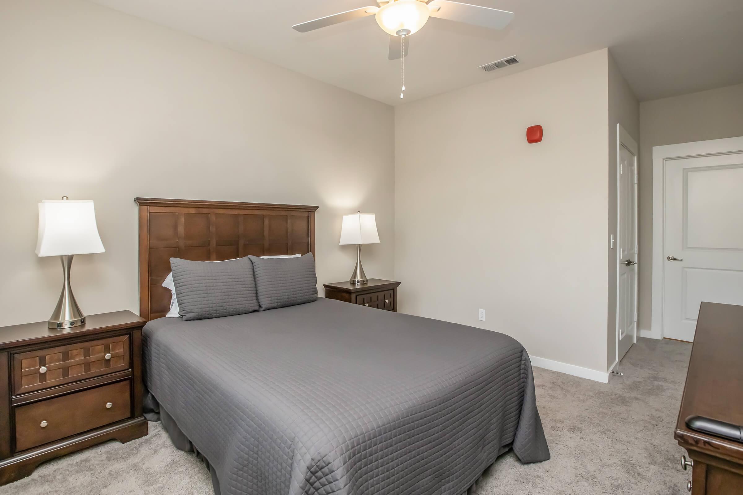 A neatly arranged bedroom features a queen-sized bed with a gray quilt and two bedside lamps on wooden nightstands. A dresser is positioned against one wall, and a ceiling fan hangs above. The room has neutral-colored walls and carpet, creating a calm atmosphere.