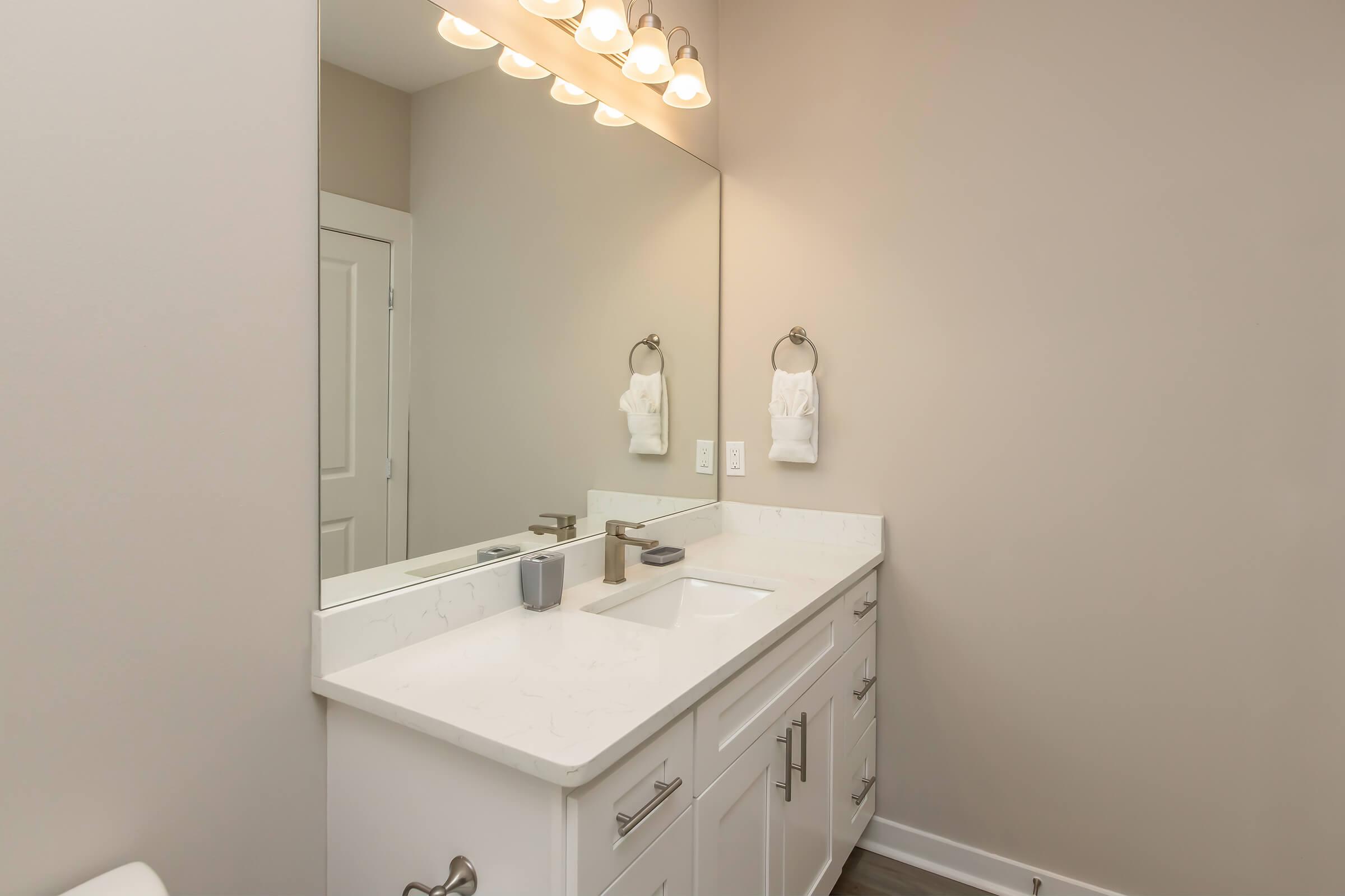 A modern bathroom featuring a double sink vanity with a white countertop, a large mirror above, and two towel holders. The walls are painted in a soft neutral color, and the lighting is provided by three overhead fixtures. The floor is a warm wood finish, adding a cozy touch to the space.
