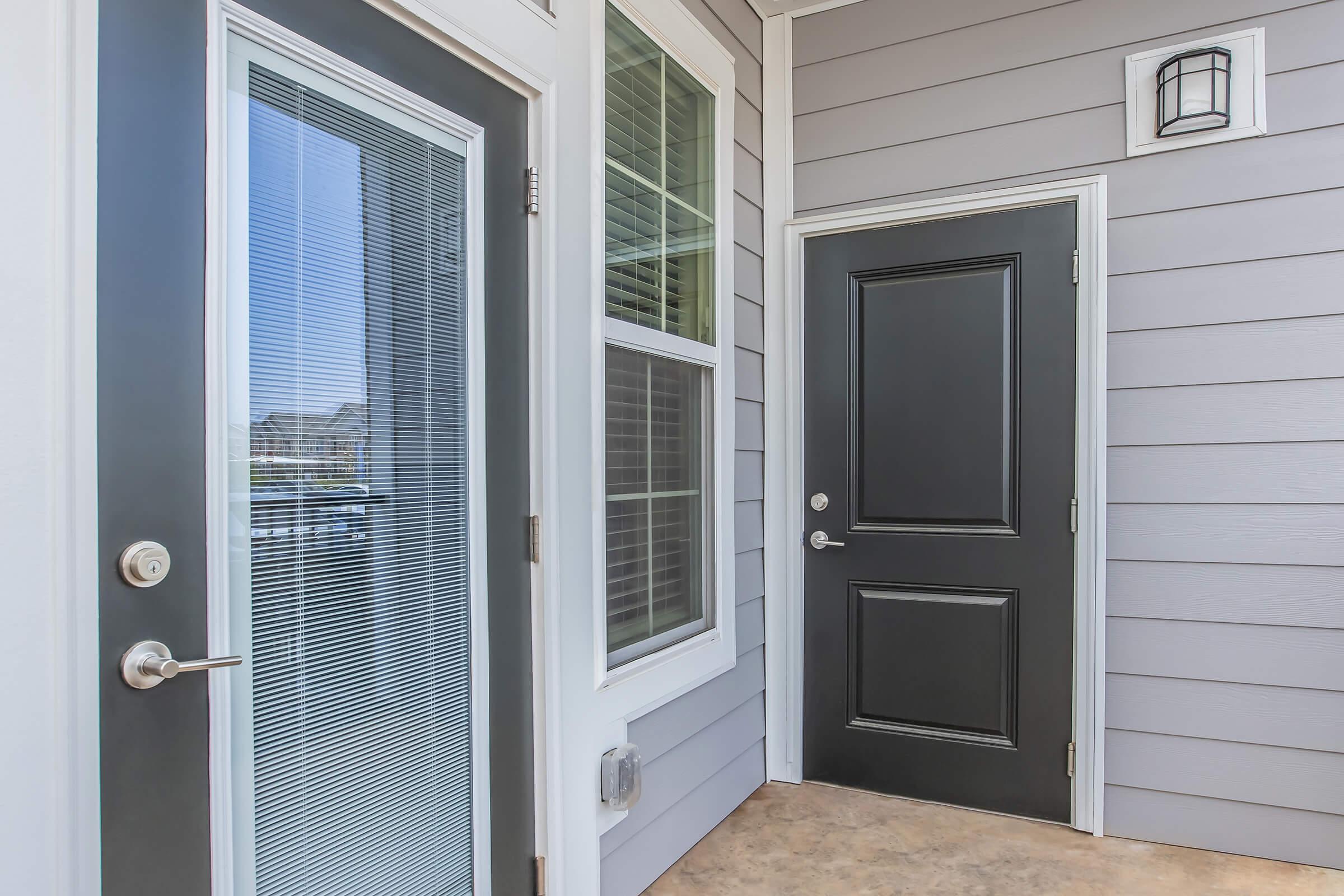 A modern entrance featuring a dark front door and a glass-panel door beside it. The walls are covered in light gray siding, and there’s a decorative light fixture above the door. The floor is tiled, and the setting appears bright and welcoming with natural light coming through the glass door.