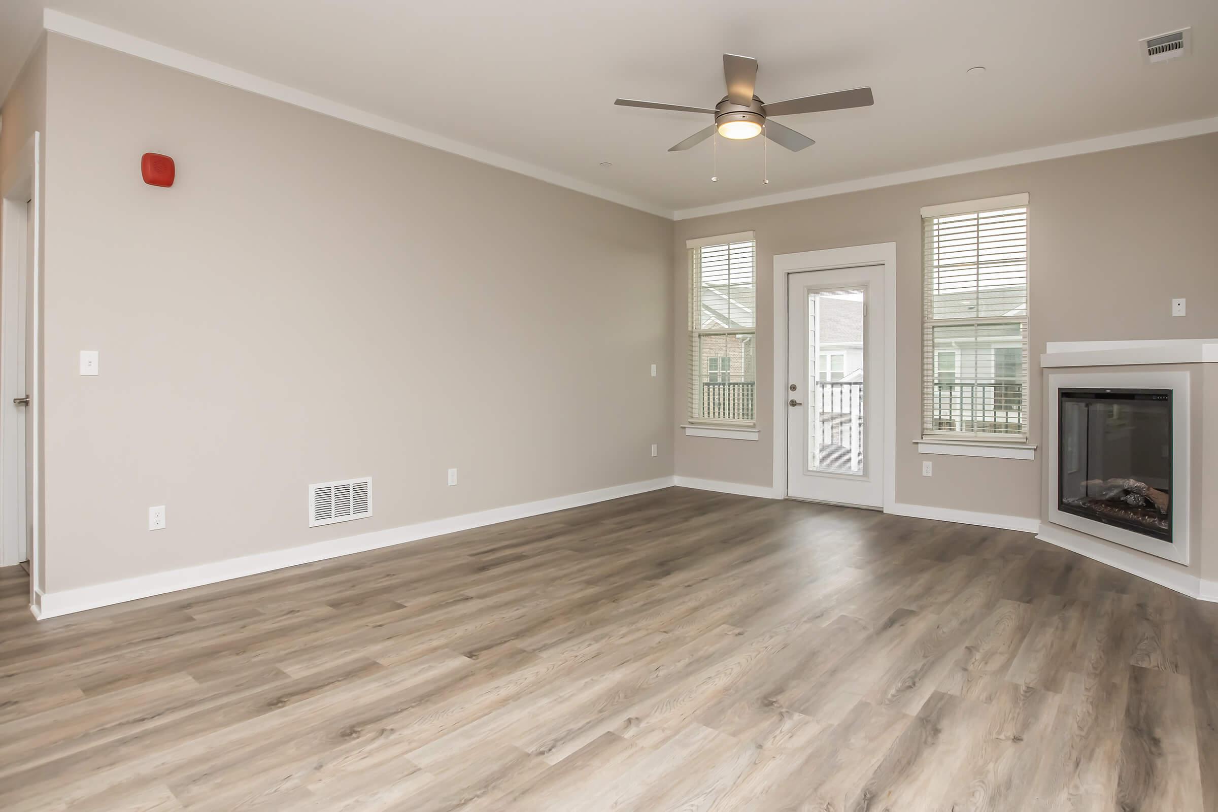 A spacious, empty living room featuring light-colored walls, modern wood flooring, and a ceiling fan. There are two large windows providing natural light and a door leading to a balcony. A contemporary fireplace is positioned against one wall, enhancing the room's cozy atmosphere.
