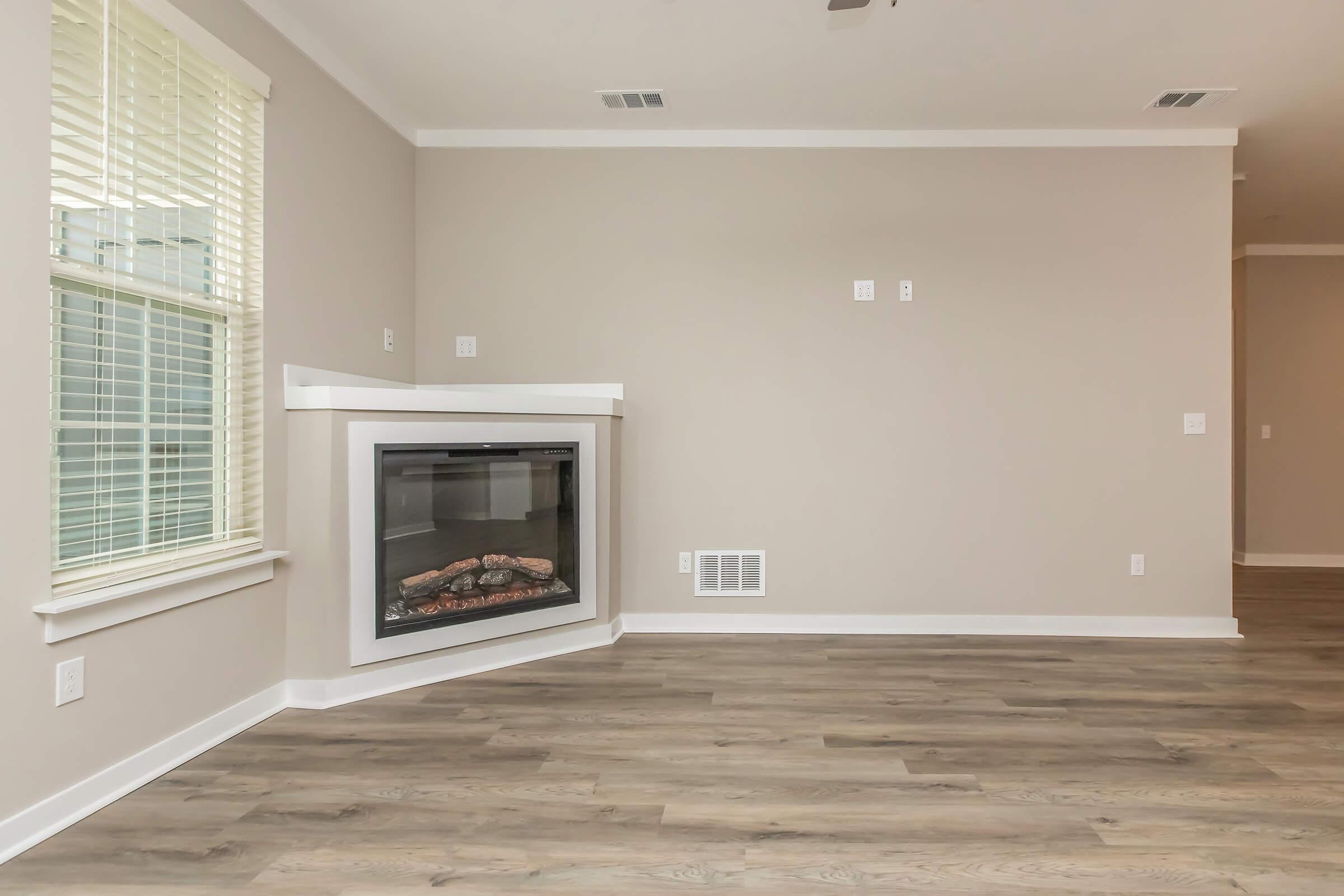 A spacious, empty room featuring light-colored walls and a modern fireplace with a glass front. The floor is made of wood laminate, and there's a large window providing natural light. The room has a clean and minimalistic design, suitable for various interior styles.