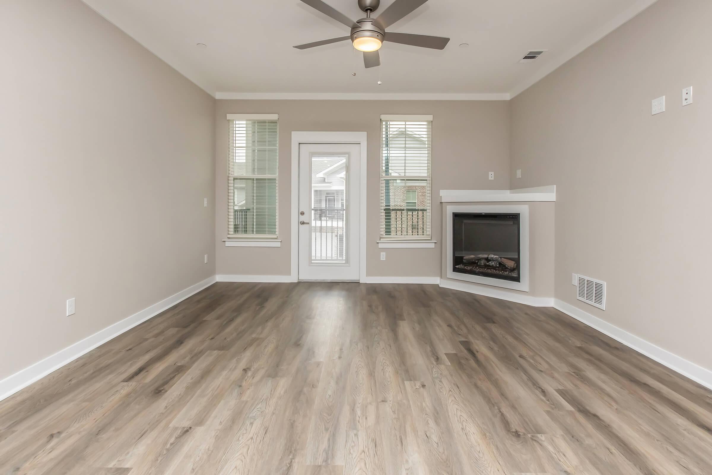 A spacious living room with light brown walls and wooden flooring. It features a ceiling fan, large windows with blinds, and a door leading outside. There is a modern electric fireplace in the corner, creating a cozy atmosphere. The room is empty, allowing for personalization.