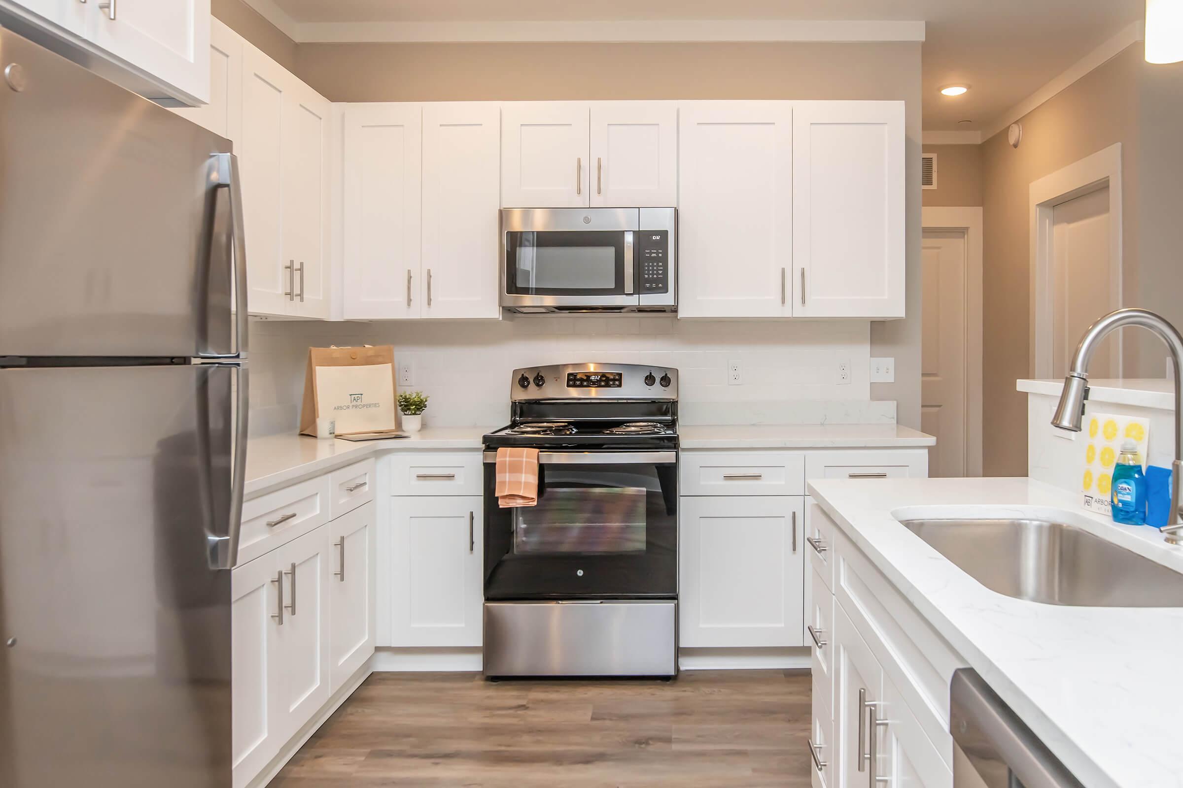 Modern kitchen featuring white cabinetry, stainless steel appliances including a refrigerator, microwave, and oven. The countertop is light-colored with a sink visible. A decorative towel hangs on the oven door, and there is a small plant on the counter. The flooring is a dark wood finish, creating a contemporary aesthetic.