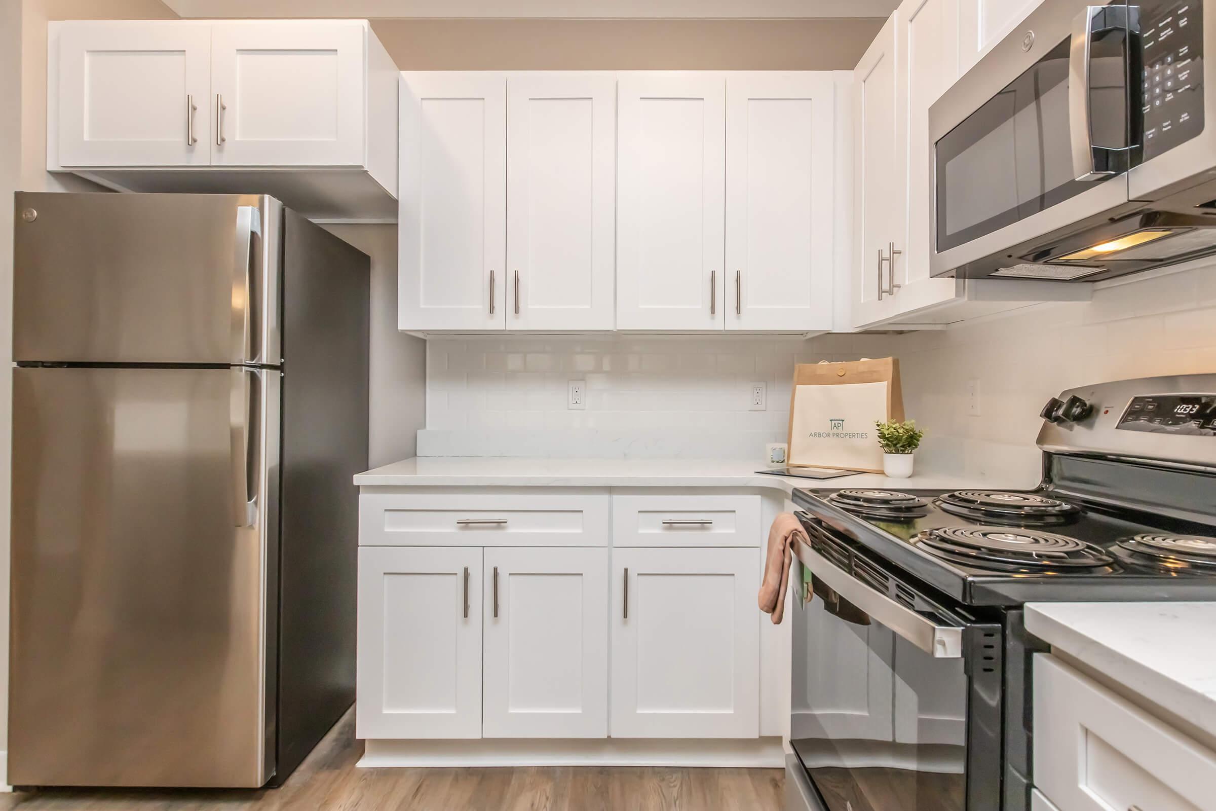A modern kitchen featuring white cabinetry, stainless steel appliances including a refrigerator and microwave, and a black stovetop. The countertop is light-colored with a small plant and a bag placed nearby. The flooring is a light wood finish, creating a bright and inviting space.