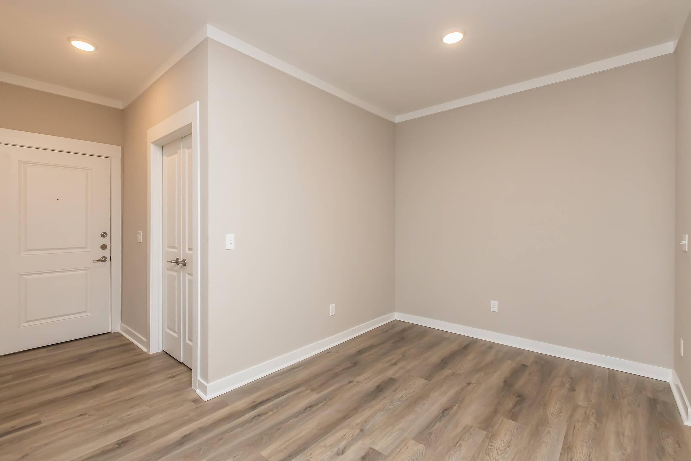 A vacant room with light gray walls and laminate flooring. There is a door on the left leading outside and a doorway on the right. The ceiling is white with recessed lighting, creating a bright and open atmosphere. The space is unfurnished, showcasing a clean and modern design.
