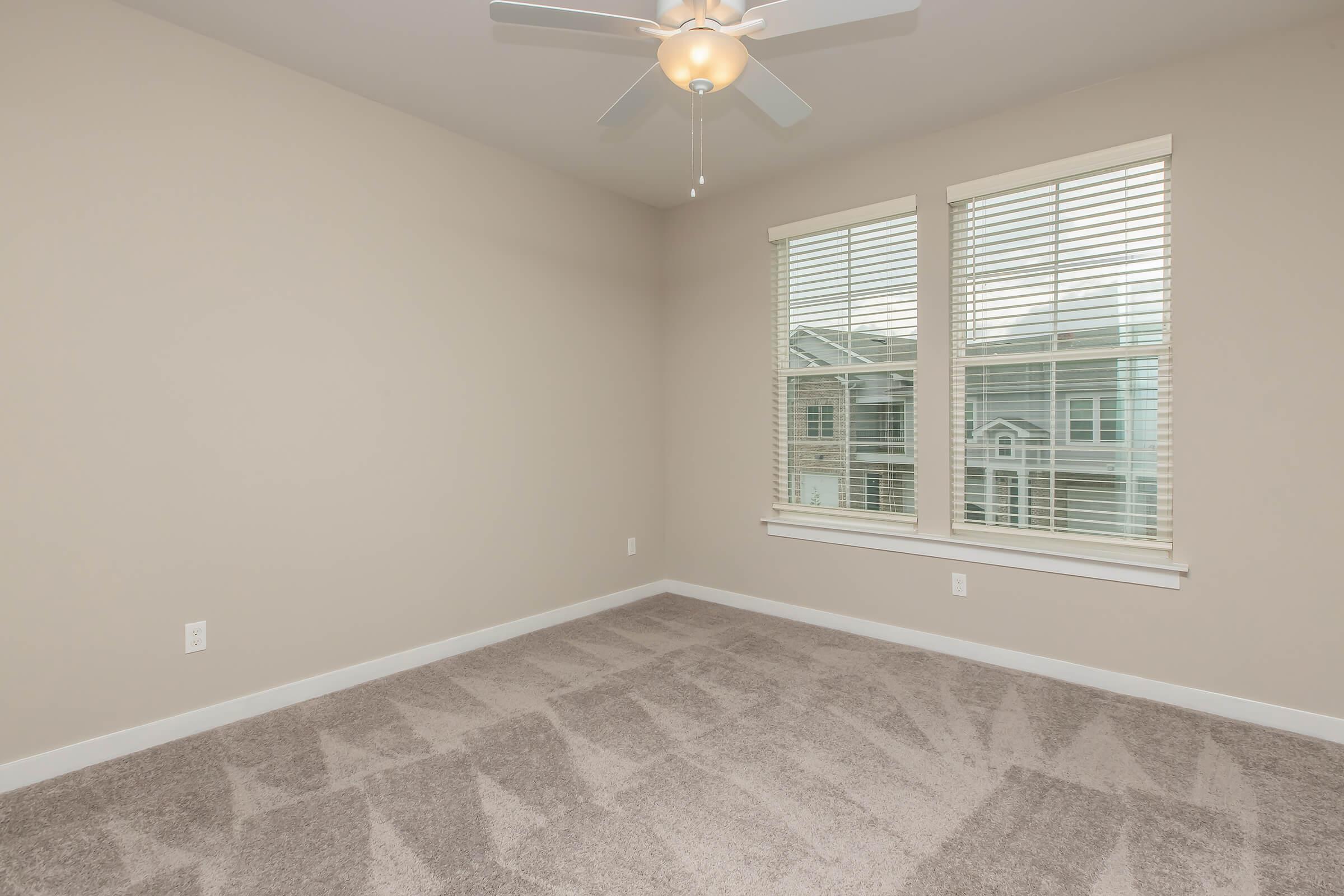 Empty room with beige walls and light-colored carpet. Two windows with blinds allow natural light to enter. A ceiling fan is installed in the center of the room, providing ventilation. The space is clean and modern, designed for versatile use.