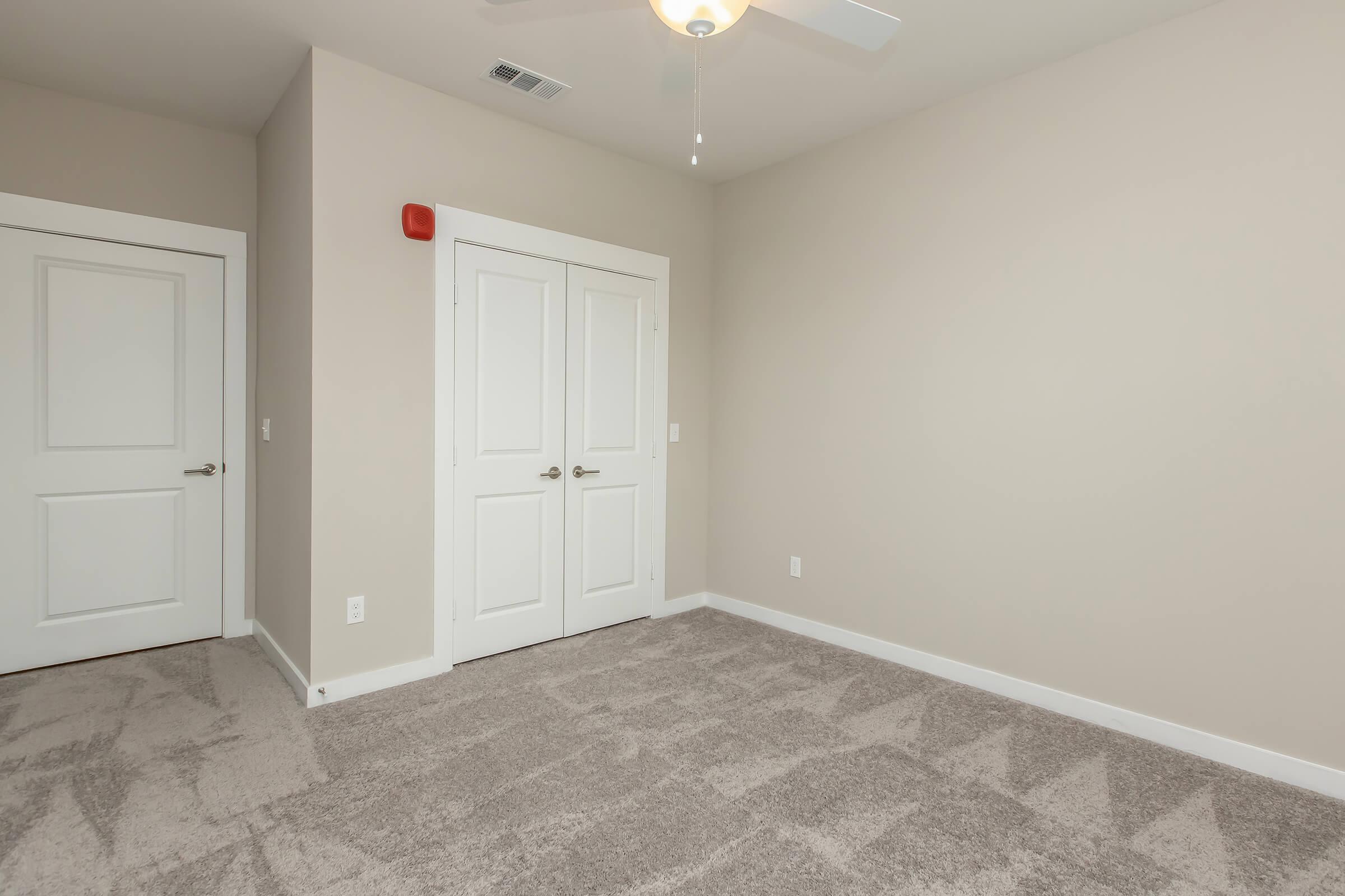 Empty room featuring beige walls and carpet, white ceiling with a ceiling fan, and a double door closet. A red fire alarm is mounted on the wall near the ceiling. The room has minimal decor and natural light intake, with one closed door visible leading to a presumably adjacent area.
