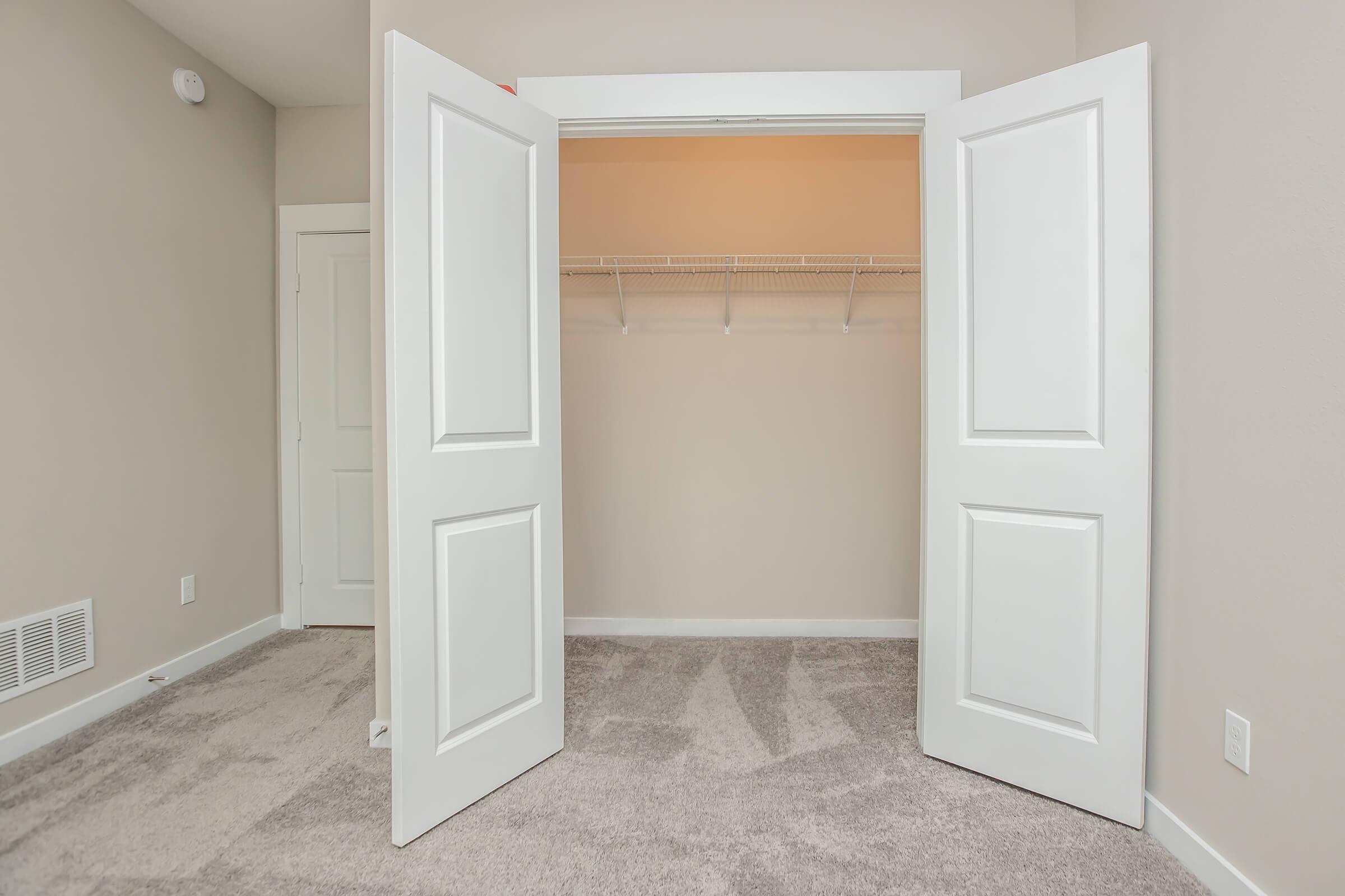 A spacious, empty closet with two open white doors, revealing a clean interior with a single shelf and beige walls. The floor is covered in light-colored carpet. There's a glimpse of another door in the background, and the overall lighting is bright and neutral.
