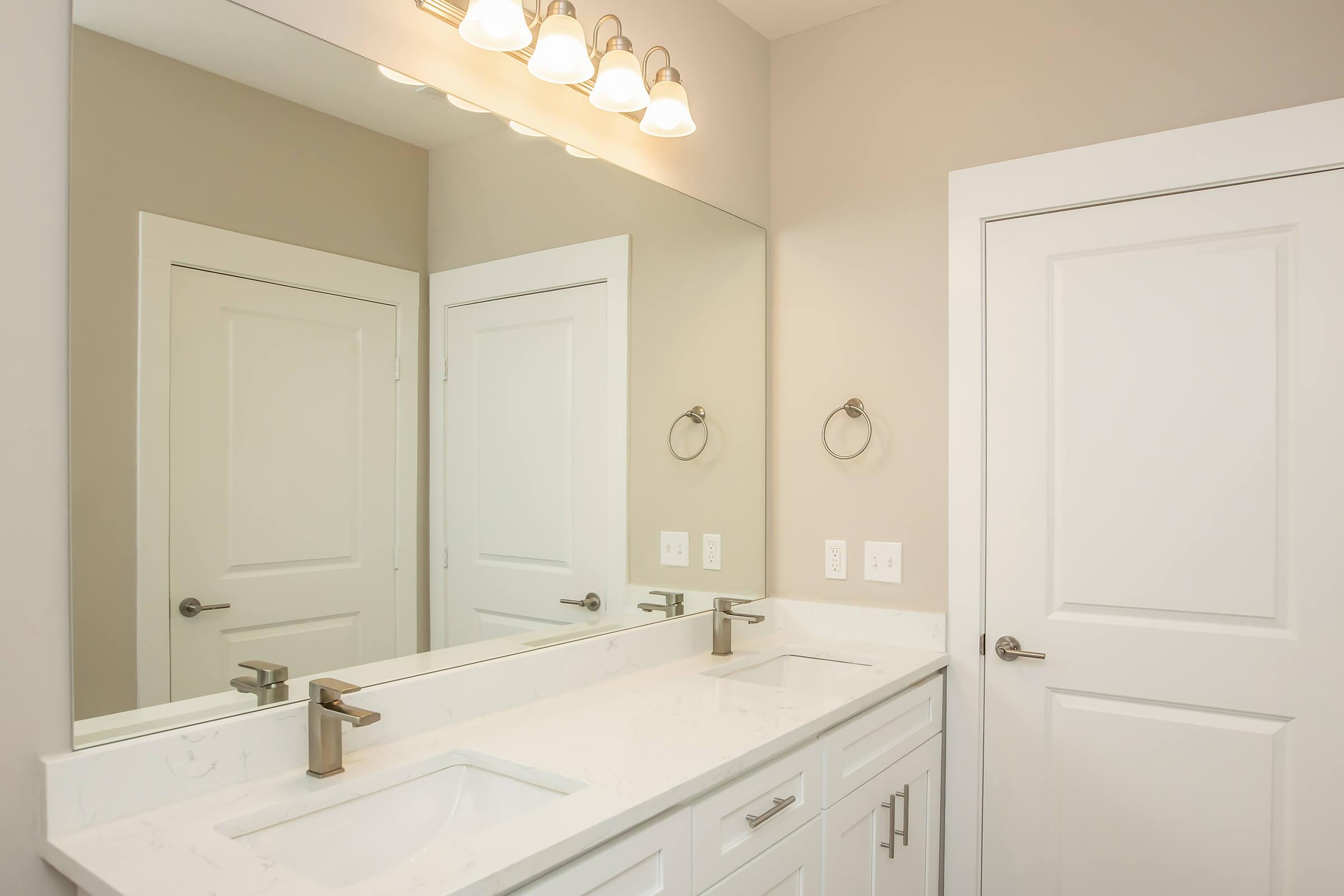 A modern bathroom featuring a double vanity with a white countertop, sleek faucets, and a large mirror above. The walls are painted in a neutral color, complemented by bright overhead lights. A white door leads to another room, and there's a towel ring on the wall beside the sink.