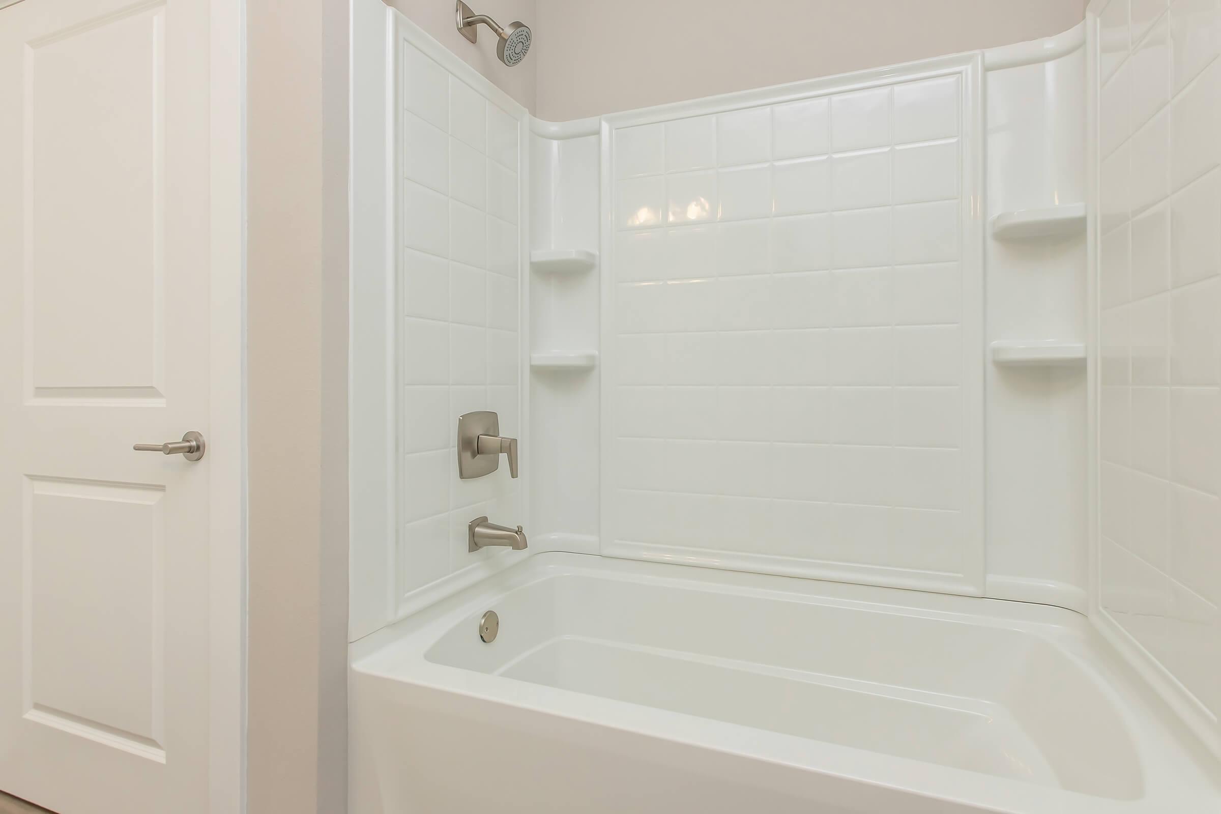 A clean, modern bathroom featuring a white bathtub with a tiled surround. The space includes two built-in shelves for storage and a showerhead above. A simple door exits the room, contributing to a minimalist aesthetic.