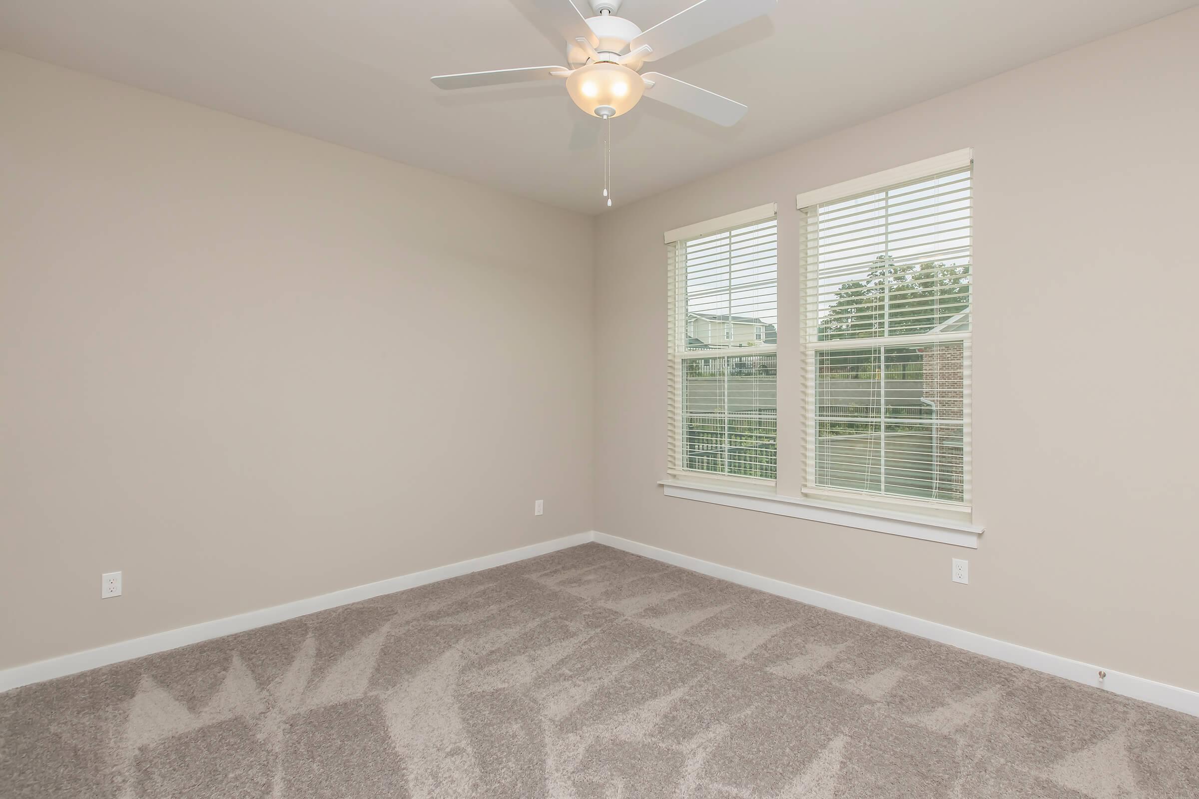 A neutral-toned room with light beige walls and a light-colored ceiling fan. It features two large windows with white blinds, allowing natural light to enter. The floor is covered with plush gray carpet, and the overall ambiance is bright and spacious, suitable for a living area or bedroom.