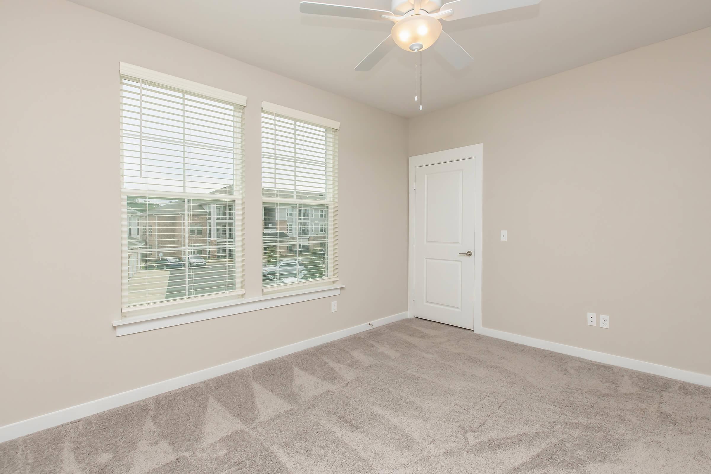 Empty room with light gray carpet and beige walls, featuring a ceiling fan and two large windows with white blinds. A white door is visible on one wall, and there is a view of an outdoor area through the windows. The space is bright and inviting, ideal for various uses.