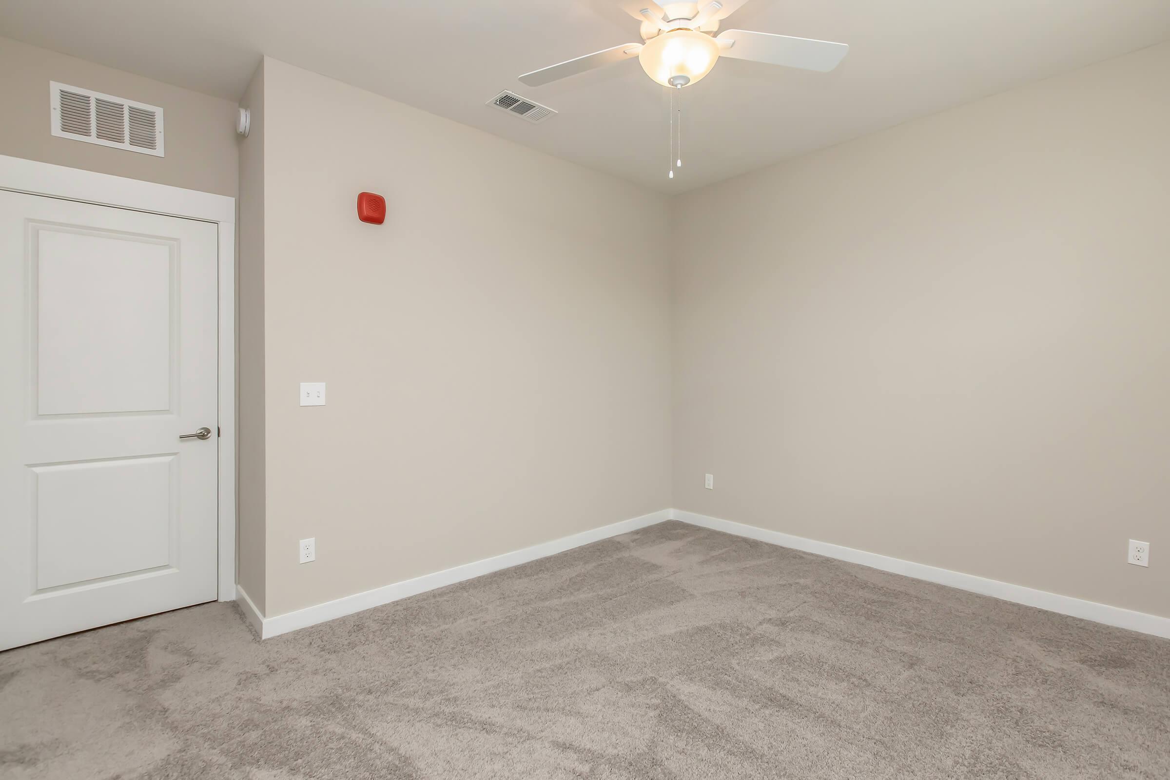 A sparsely furnished room with light beige walls and a ceiling fan. The space features a door on the left, a smoke detector on the wall, and soft carpet flooring. The room is well-lit, creating a clean and inviting atmosphere, suitable for various purposes such as an office or bedroom.