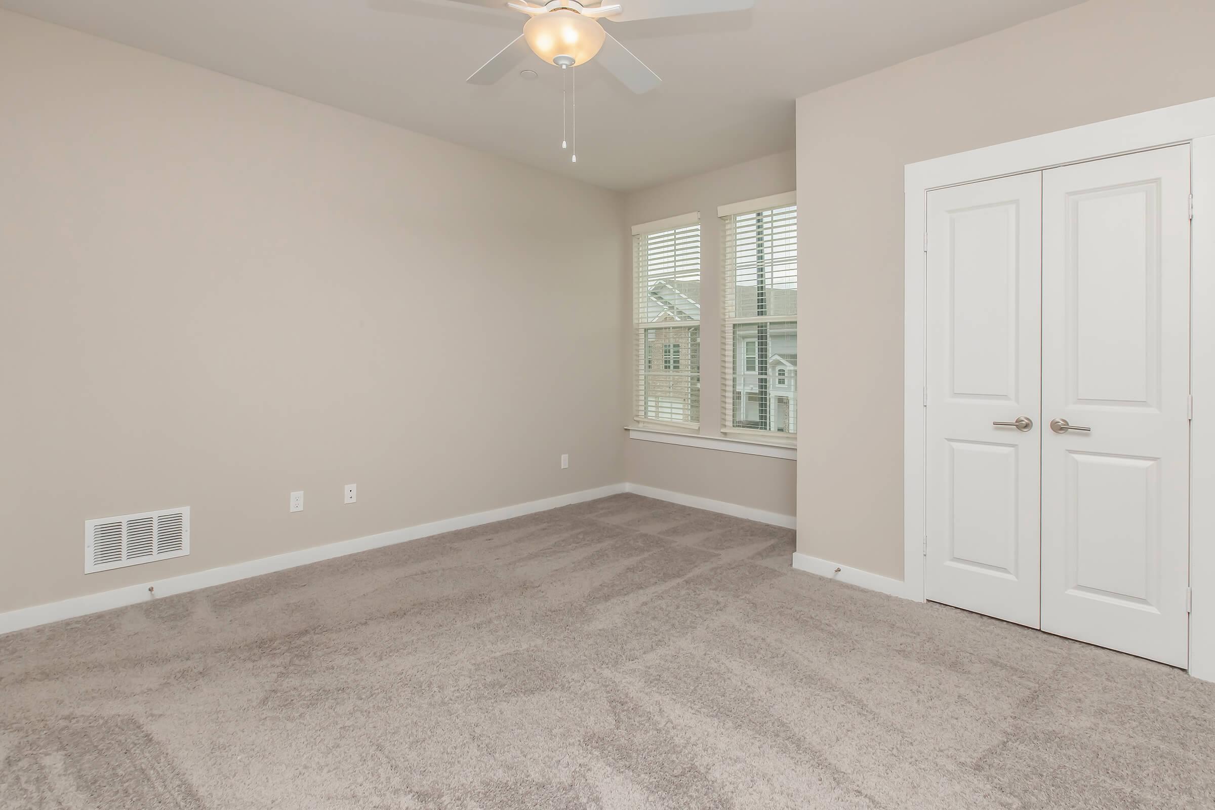 Interior of a small, empty bedroom featuring light beige walls, a ceiling fan, and window with white shutters. The floor is carpeted in a soft, neutral shade. There is a double-door closet on the right side, and the overall atmosphere is bright and inviting.