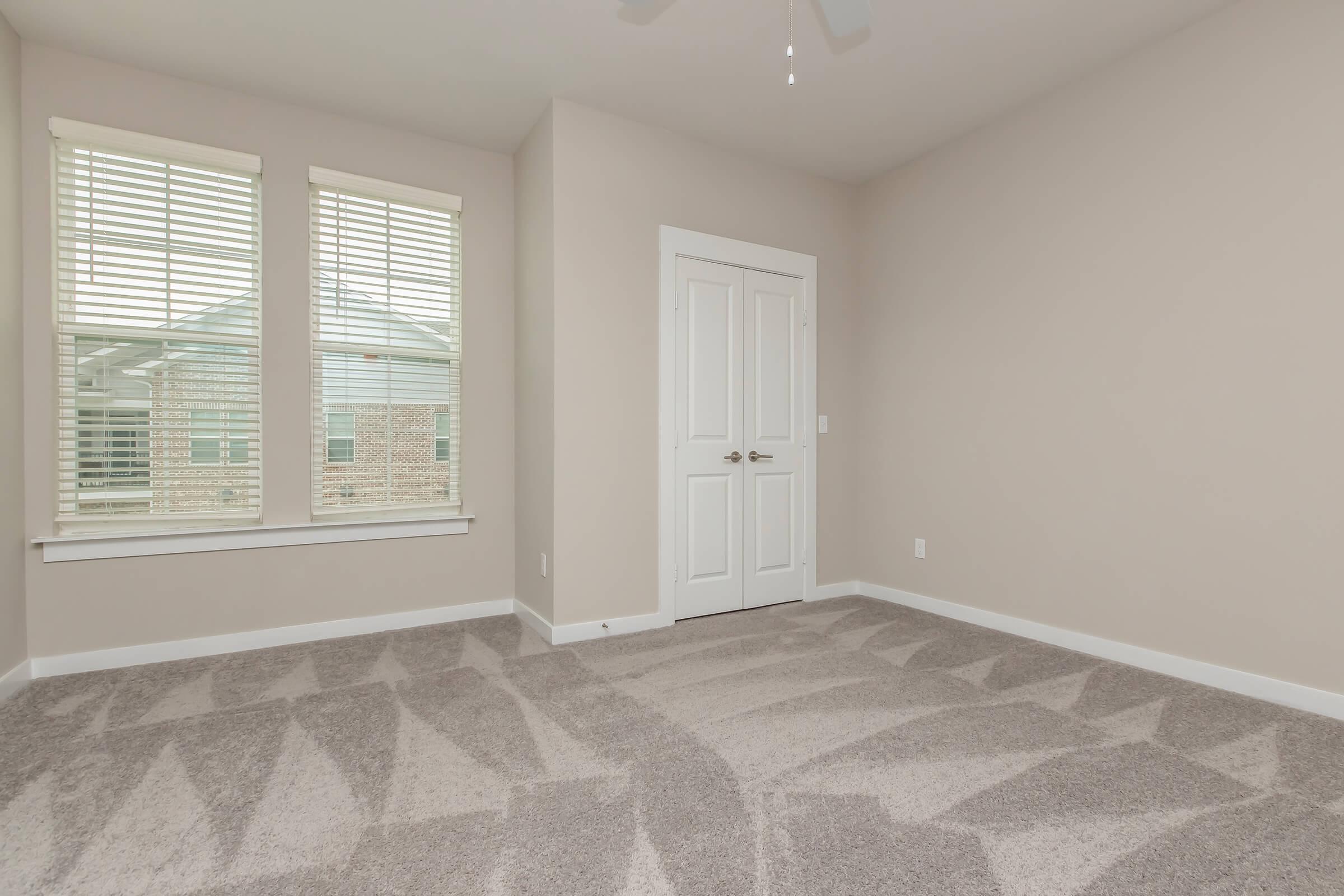 A clean, empty room with light beige walls, a ceiling fan, and two windows with white blinds letting in natural light. The floor features light gray carpeting with a subtle pattern. A white door with a silver handle leads to an adjacent space. The overall atmosphere is bright and spacious, ready for furnishing.