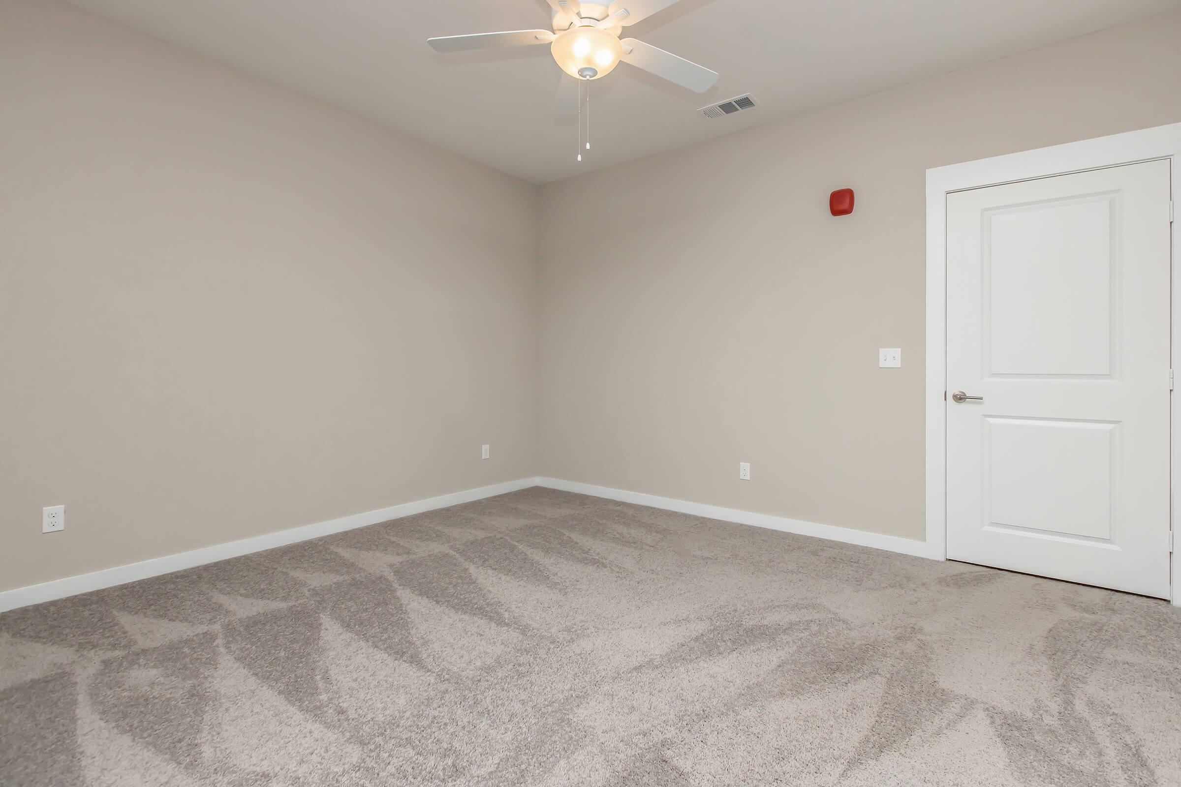 Empty interior room with light beige walls and plush gray carpet. A ceiling fan is mounted in the center, and there's a white door on the right side leading to another area. A small red fire alarm is visible on the wall, adding to the minimalistic and clean design of the space.