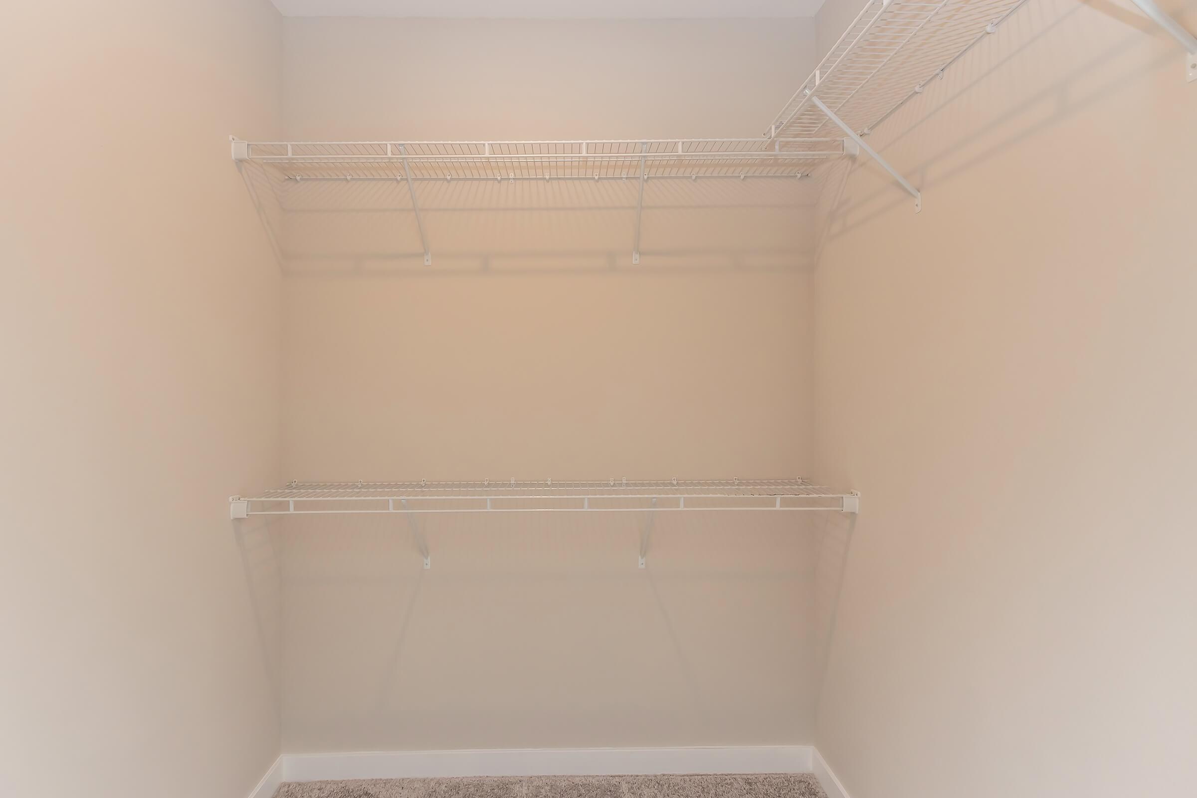 A clean, empty closet with white wire shelving against a light beige wall. The carpeted floor is visible, and there are no items stored inside. The shelving consists of two horizontal units with additional vertical supports.