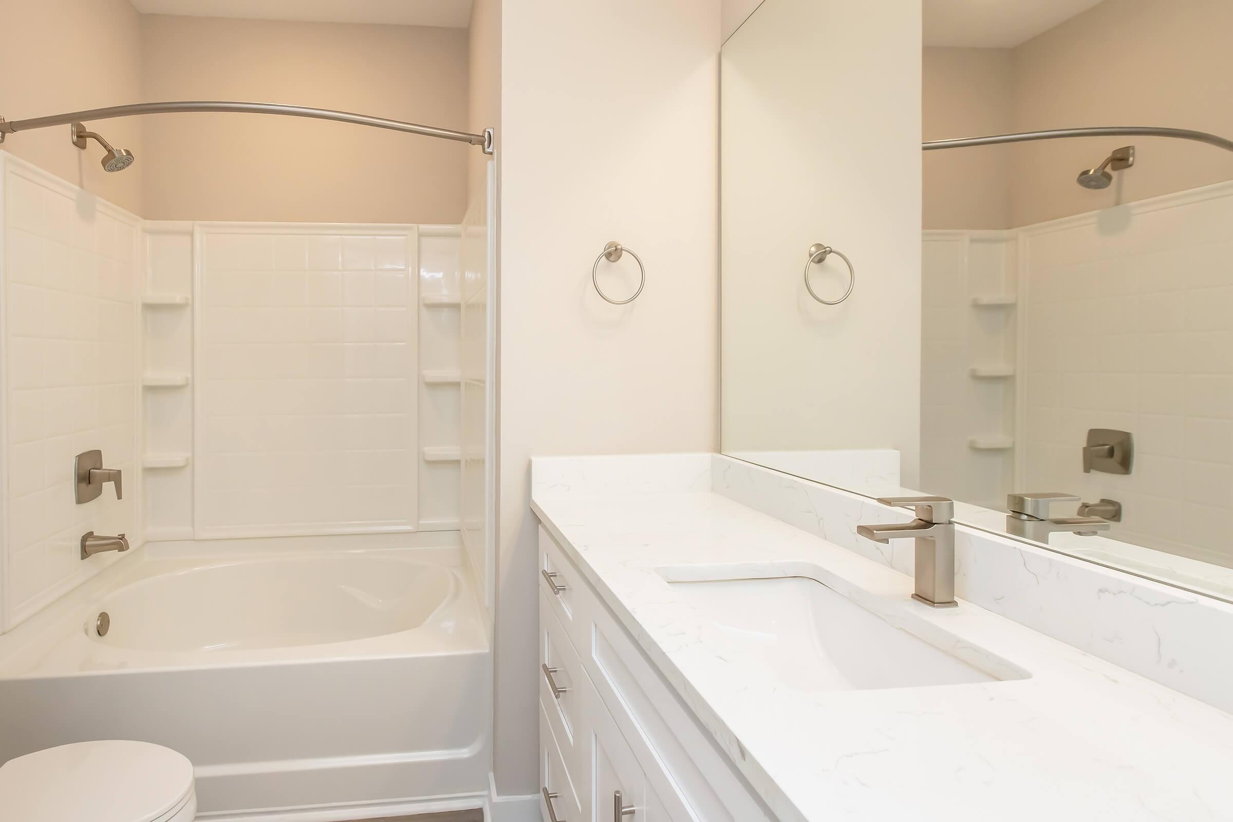 A modern bathroom featuring a white bathtub with a shower, light-colored walls, a sleek sink with a marble countertop, and a large mirror. The space is well-lit and includes silver towel rings and modern fixtures, creating a clean and refreshing atmosphere.