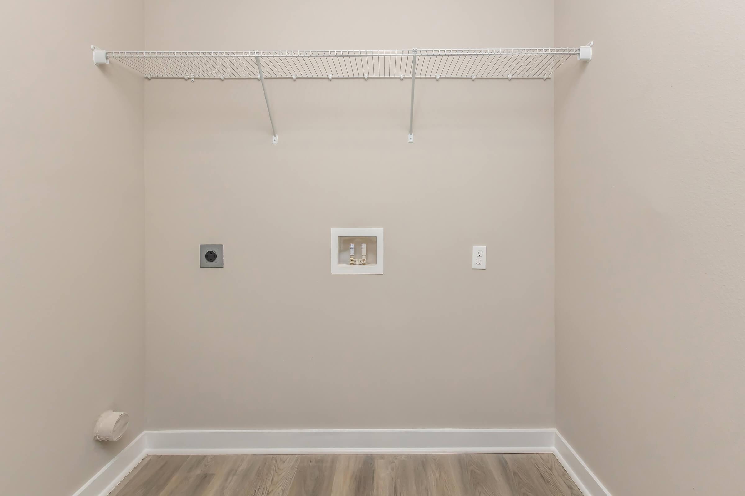 Empty laundry room with plain beige walls. A wire shelf runs along the back wall, and there are utility connections for a washer and dryer. The floor is wood-like laminate, and there are no additional items or decorations in the space.