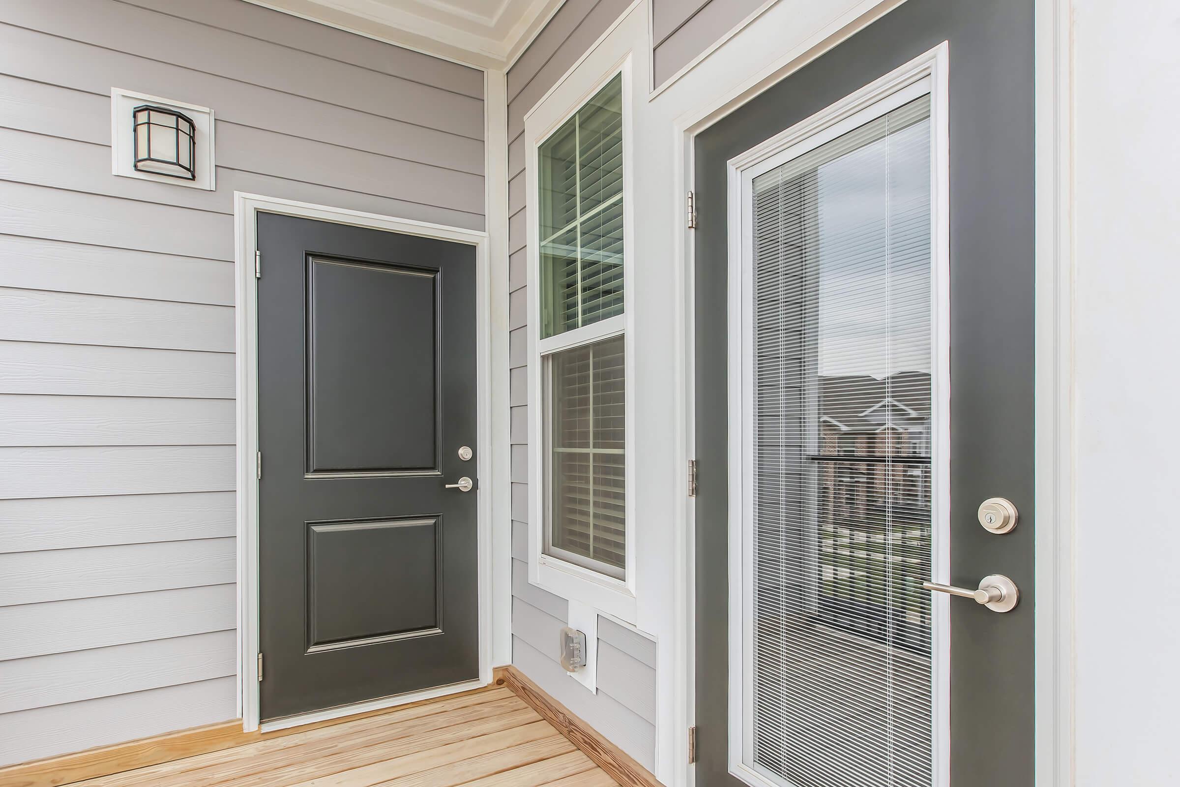 A welcoming entrance featuring a dark front door and a glass-panel door next to it, both set against light gray siding. The area has a wooden floor and a modern light fixture above, providing a bright and inviting atmosphere for visitors.